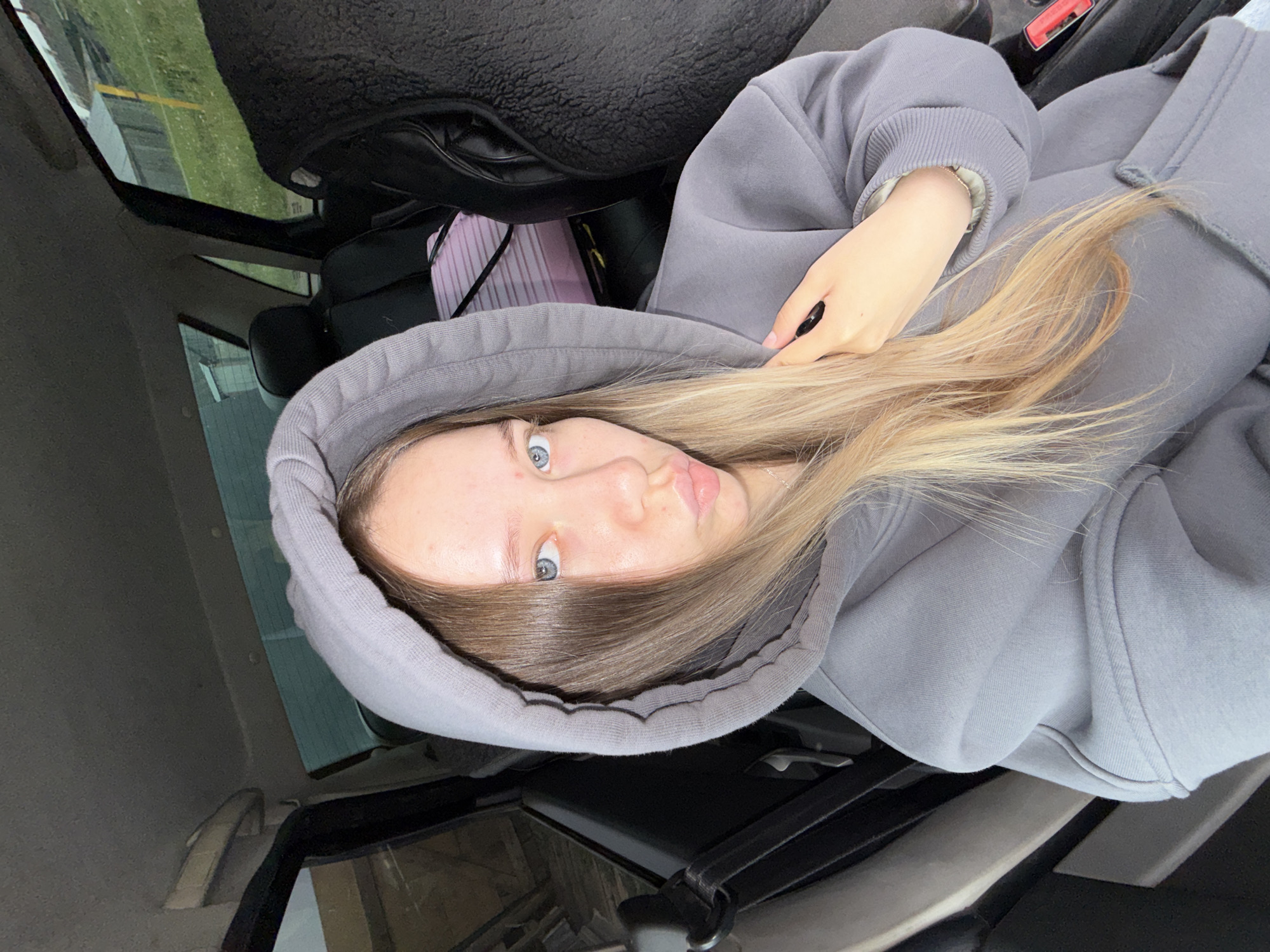 A young woman with long, straight blonde hair and blue eyes is the central subject of the image, capturing a selfie inside a car. She is wearing a grey oversized hooded sweatshirt, with the hood pulled up around her head. She looks directly at the camera with a slight pout on her lips. Her fair skin shows a few minor blemishes on her forehead. A silver necklace chain is visible around her neck, and her right hand, with black nail polish on her nails, is partially visible, holding the phone she is using to take the picture.
The setting is the interior of a car, likely from the passenger perspective. Dark-toned car seats and the car's roof are visible. Through the car window in the upper left, a green grassy area and what appears to be a building or distant structure are visible under an overcast sky, indicating daytime. A red seatbelt buckle with some unreadable white text is visible to the woman's right. A pink striped item, possibly a bag, is tucked beside the seat. The overall scene conveys a casual and relaxed moment, possibly taken while traveling or waiting in a car in Kamennyi Kliuch, Russia. No clearly legible text is visible within the image.