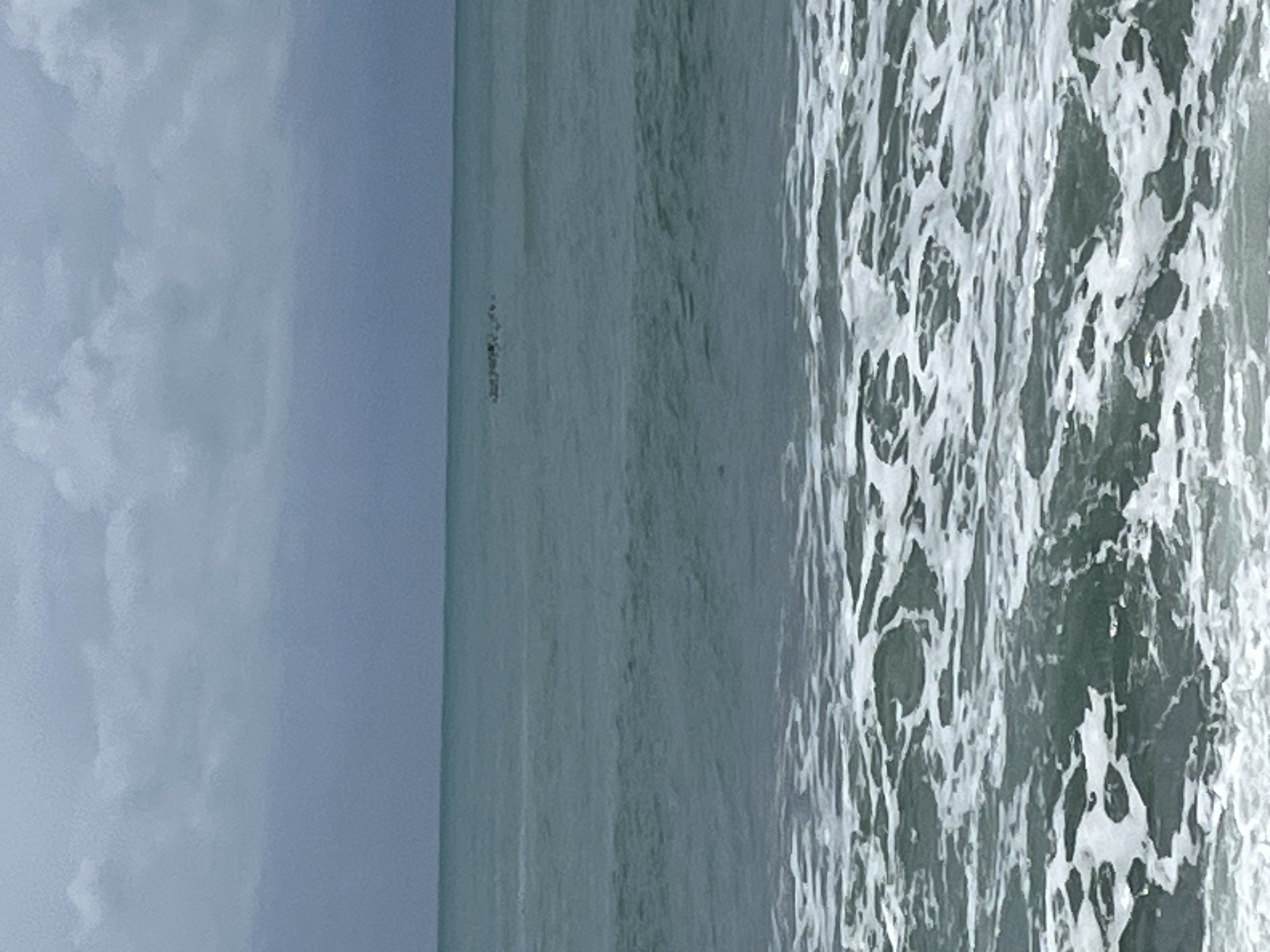 The image depicts a seascape with a gray sky on the left side of the image. The ocean occupies the majority of the central portion of the image, transitioning from relatively calm to choppy waters with white foam on the right side. The location is possibly near the city of Alanya, Turkey. However, without additional context, it is hard to describe the exact geographical location from this picture. There are no people visible in the image.