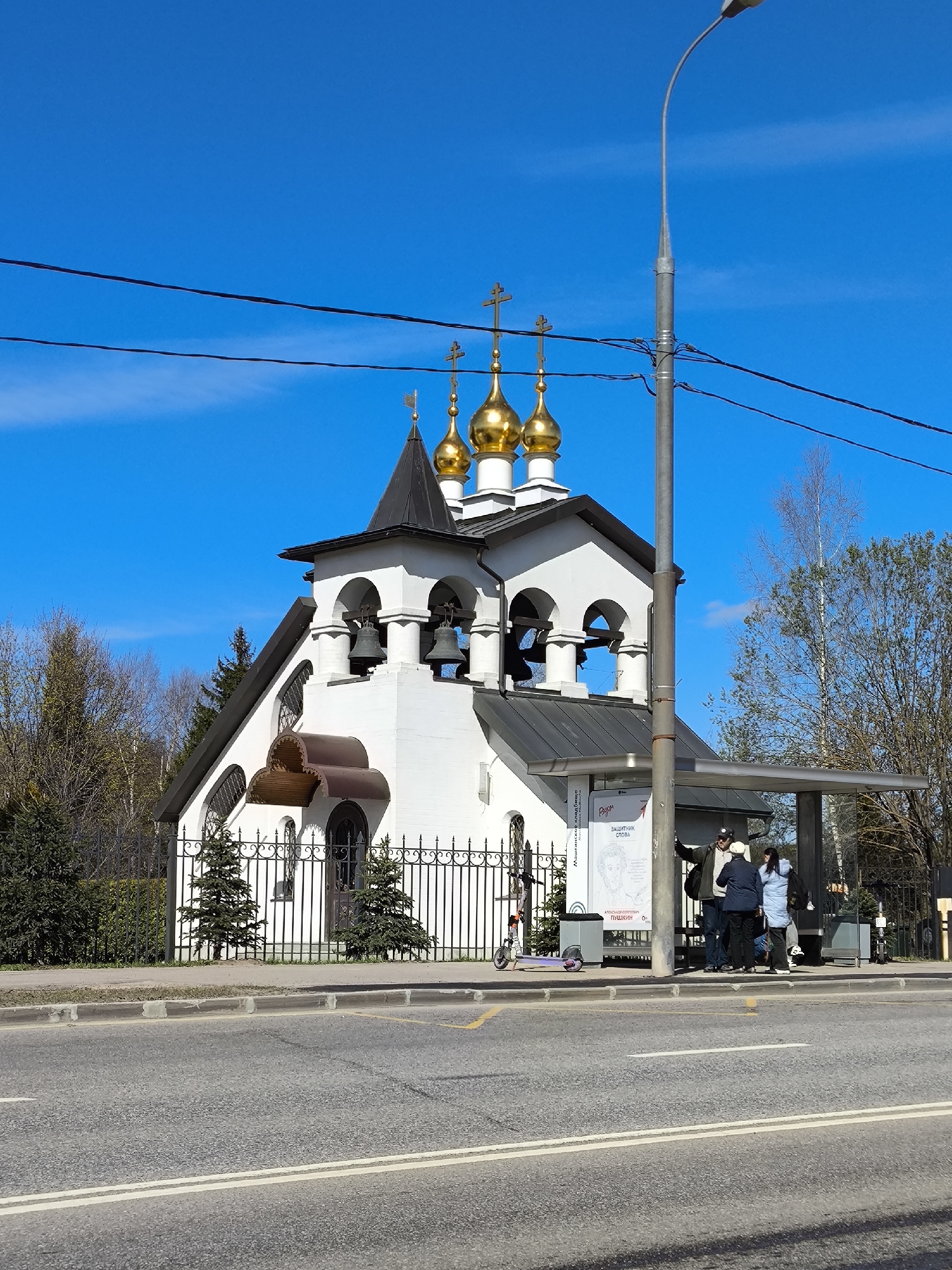 The image shows a white, ornately-decorated church with golden domes, likely an Orthodox church, located on a street in Moscow, Russia. A bus stop adjoins the church, where a group of people are waiting. A light pole stands to the right of the church and the bus stop. A fence surrounds the church. A scooter is parked near the bus stop. A road with white lines runs in front of the church and bus stop. The sky is clear and blue. Trees are visible in the background.