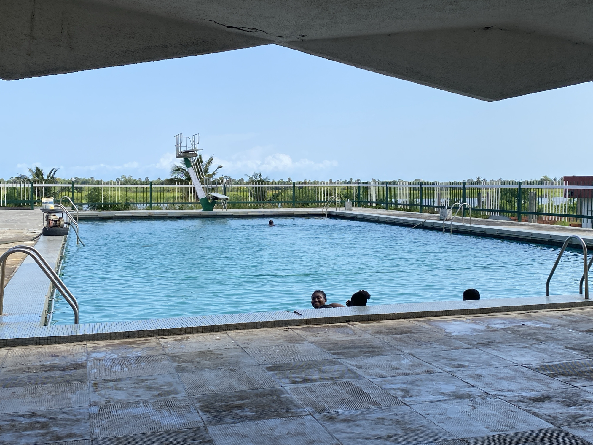 The image shows an outdoor swimming pool in Tawpo, Nigeria. The pool is rectangular and filled with clear blue water. Several people are in the pool, with at least two visible near the foreground edge. One person is submerged with only the top of their head visible. A diving board is present at the far end of the pool, with a white railing lining the pool's perimeter, backed by green bushes. The pool is surrounded by grey tiled flooring. An overhead concrete structure partially frames the view. The sky is clear with a light blue hue.