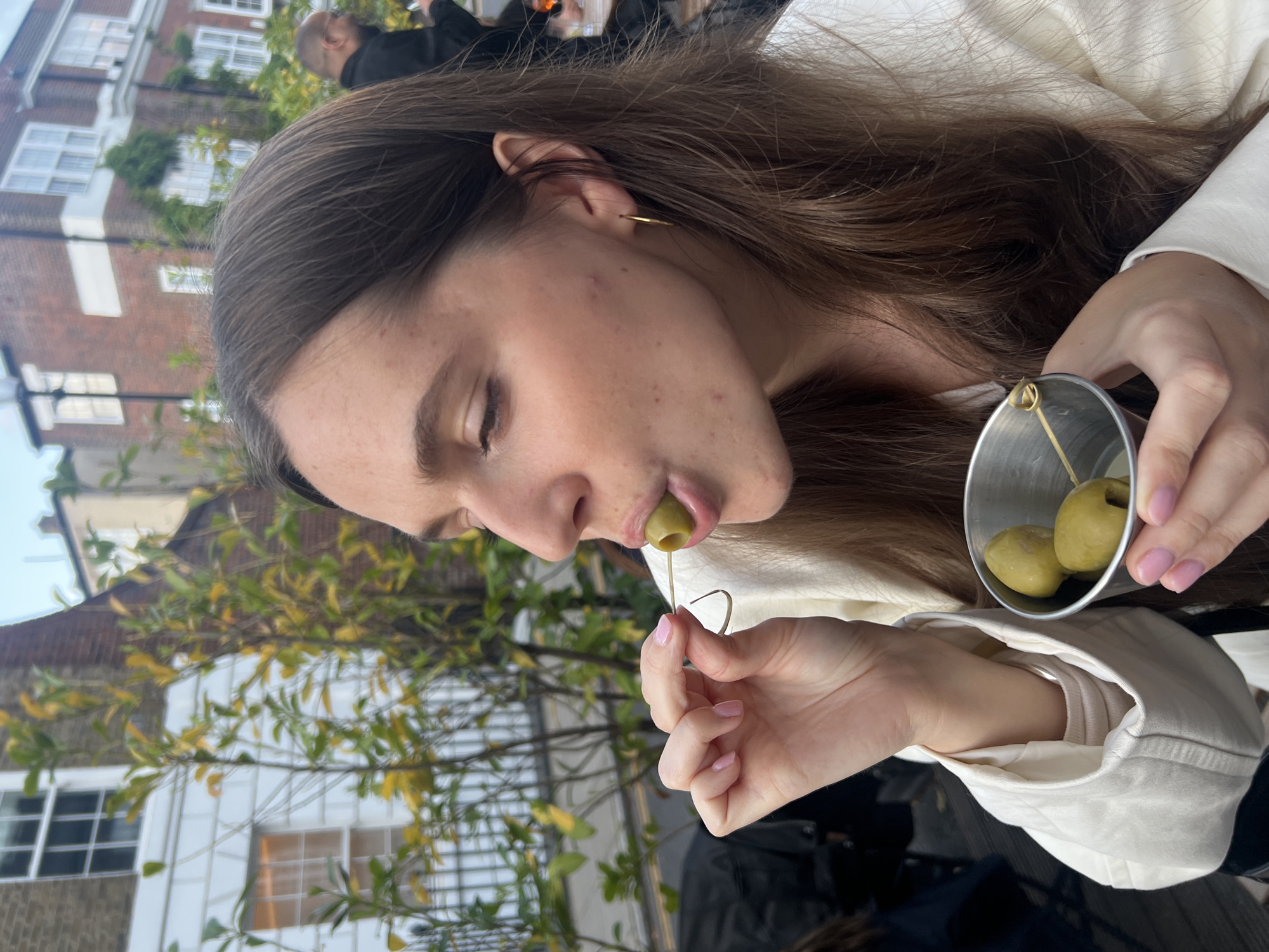A woman is about to eat a green olive impaled on a cocktail stick. She is holding the olive in her right hand and a small, silver bucket containing two more olives in her left. She has long brown hair, a gold hoop earring, and her fingernails are painted light pink. She is wearing a white shirt or jacket. The background shows a building, possibly a house, constructed of red brick and white painted window frames. There are green trees and foliage. The picture was likely taken outdoors, in a city environment. The location appears to be in London, United Kingdom.