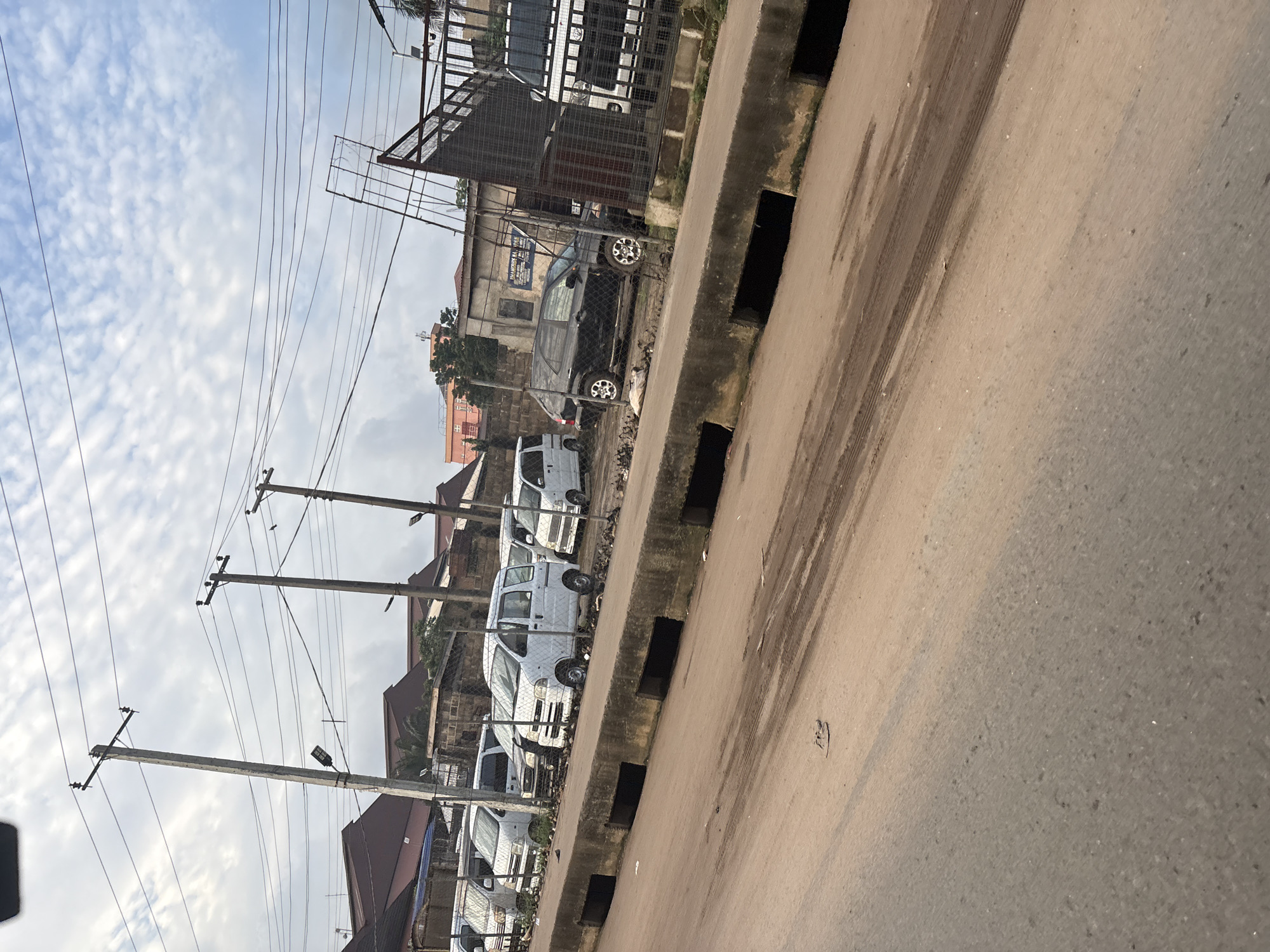 The image captures a view of a street in Onitsha, Nigeria, characterized by a row of parked white commercial vans and a black SUV. The vehicles are positioned behind a low concrete barrier with dark, rectangular openings, suggesting a drainage system. A wire fence separates the vehicles from a building in the background, which appears to be a commercial establishment with a sign that is partially visible. Overhead, a network of power lines and utility poles dominate the sky, which is partly cloudy. The foreground shows a dusty, reddish-brown road with tire tracks, indicating recent vehicle activity. The overall scene depicts a typical urban streetscape in Nigeria, with a focus on the parked vehicles and the surrounding infrastructure.