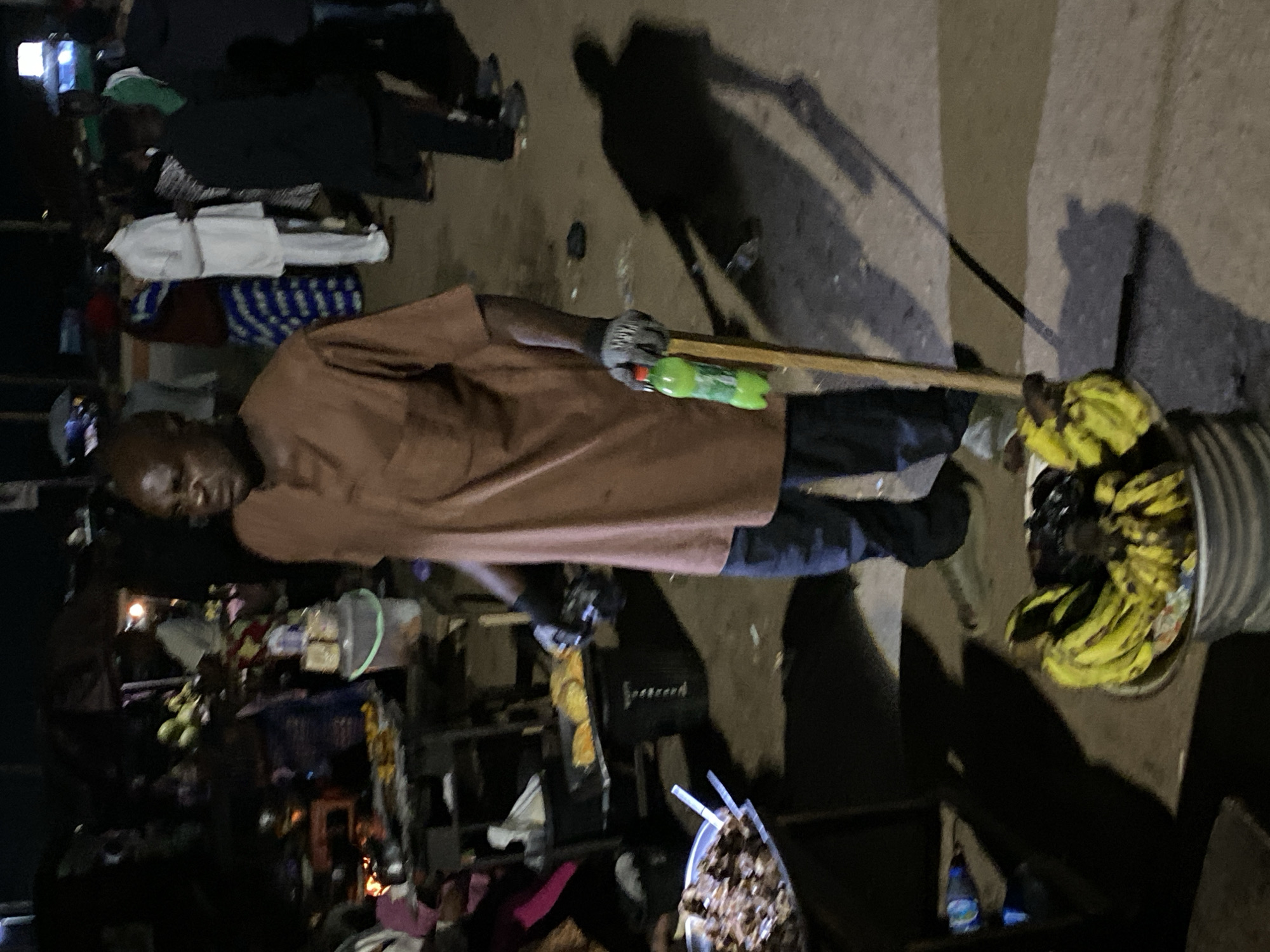 This image captures a nighttime scene in Bukuru, Nigeria, depicting an informal market or street food environment. The central figure is an adult male with dark skin and a bald head, wearing a brown long-sleeved tunic shirt and dark trousers. He is also wearing light-colored work gloves. In his right hand, he holds a green plastic bottle with a red cap, resembling a soda drink, and a vertical wooden pole. His shadow is cast prominently on the ground to his right, extending away from him.
To his lower left, a large corrugated metal bucket is visible, filled with numerous ripe yellow bananas and some darker fruit, possibly mangoes or avocados, suggesting he is a fruit vendor. Further to the left, partially obscured, is a metal pan containing what appears to be cooked meat pieces.
In the mid-ground, behind the central figure and to his left, there is a makeshift stall or table with various items. A cooking apparatus with frying food (yellowish items, possibly plantains or yams) is visible, illuminated by a light source within the stall. A black trash bin or container with white lettering is positioned below this cooking area. A white bucket with several cups or small containers on top is also present.
The background is populated with several blurred figures, indicating movement and a busy setting. Notable figures include a person in a white long-sleeved kaftan and another individual wearing a blue and white patterned skirt with a reddish-brown top. Multiple other people, some partially visible, contribute to the crowded atmosphere. The ground appears to be unpaved dirt or concrete, showing wear and tear. Artificial lighting sources, likely from the stalls and overhead, illuminate the scene, creating strong shadows and highlighting the subjects against the dark night.