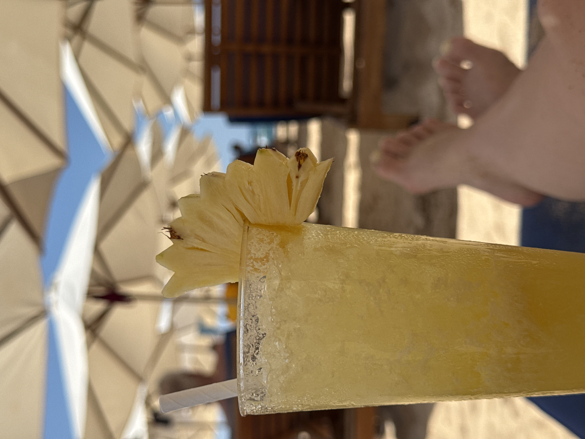 A close-up, eye-level shot focuses on a tall glass of a yellow, icy beverage garnished with a pineapple wedge. A white paper straw is visible in the drink. The glass is on a sandy surface, and in the blurred background, beach umbrellas and lounge chairs are set up. Two pairs of bare feet are in the foreground, crossed and relaxed, suggesting a leisurely beach setting. The environment appears to be a tropical beach.