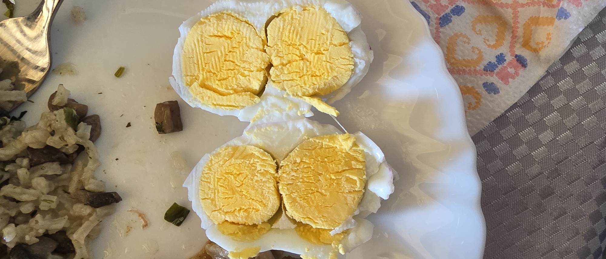 A partially eaten meal is on a white plate. The meal consists of rice with mushrooms and a hard-boiled egg cut in half. The egg yolks are visible. A silver fork is visible on the left side of the plate. The plate is on a table with a patterned tablecloth. The image was taken indoors. The location and city of Novachene, Bulgaria are not discernible from the provided image.