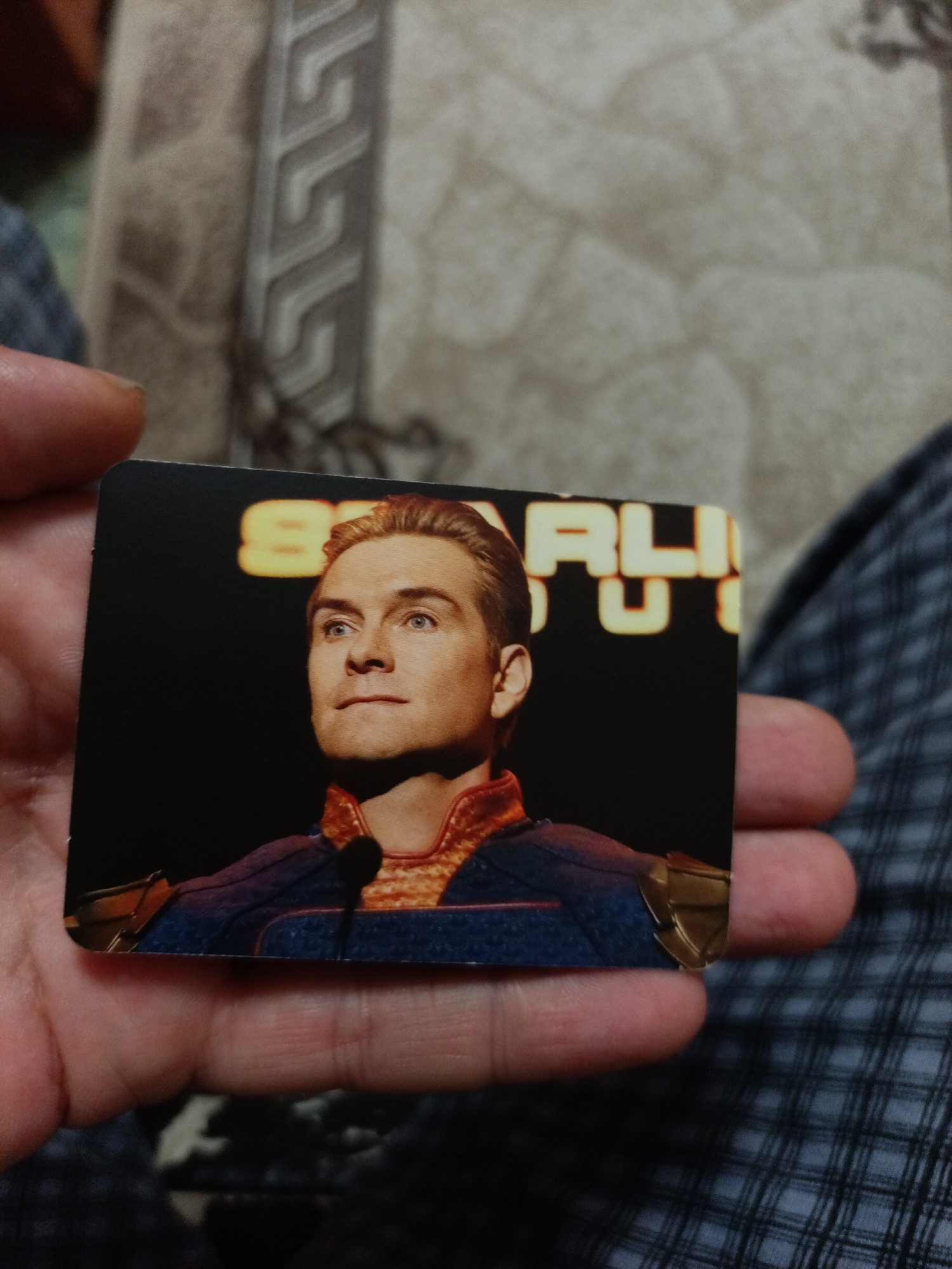 A hand holds a trading card featuring actor Antony Starr as Homelander from the TV show "The Boys". The card depicts Starr in character, looking slightly off-camera, wearing his blue and gold superhero costume. Behind him, the letters "SRLIGH" are visible in large, glowing yellow font. The background is dark. The card is held against a textured, patterned surface. The image appears to be taken in a casual setting, possibly indoors. There is no information in the image to suggest any connection to Nyagan, Russia.