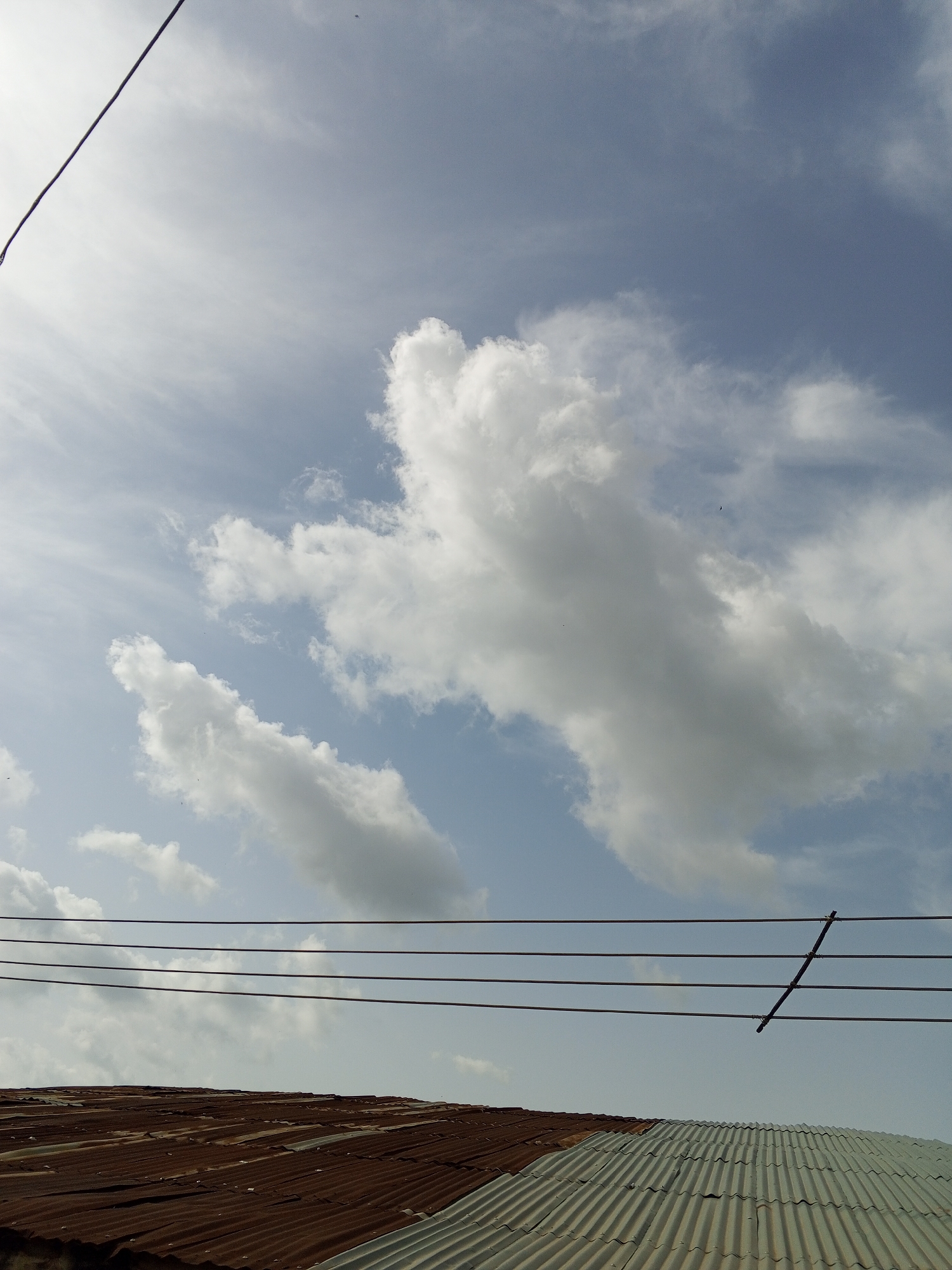 The image captures an outdoor scene in Gwong, Nigeria. In the foreground, a corrugated metal roof is visible, with a rusty brown section on the left and a silver-gray section on the right. Above the roof, four parallel electrical wires stretch across the frame, with a diagonal support structure attached to them on the right side. The background is dominated by a blue sky with scattered white clouds, including prominent cumulus clouds. A single electrical wire is also seen cutting through the upper left corner of the image.