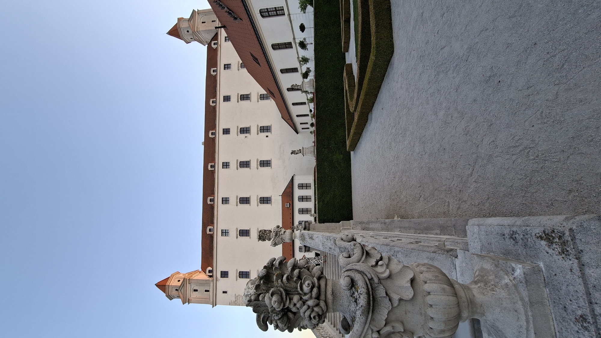 The image shows Bratislava Castle in Bratislava, Slovakia. The castle is a large, white building with red-tiled roofs and two prominent towers with conical roofs. The building is set against a clear blue sky. In the foreground, there is a stone balustrade with an ornate vase or urn. Beyond the balustrade, a pathway leads towards a manicured garden with trimmed hedges and geometric flowerbeds. There are also statues visible in the garden area. The overall scene depicts the exterior of a historical landmark.