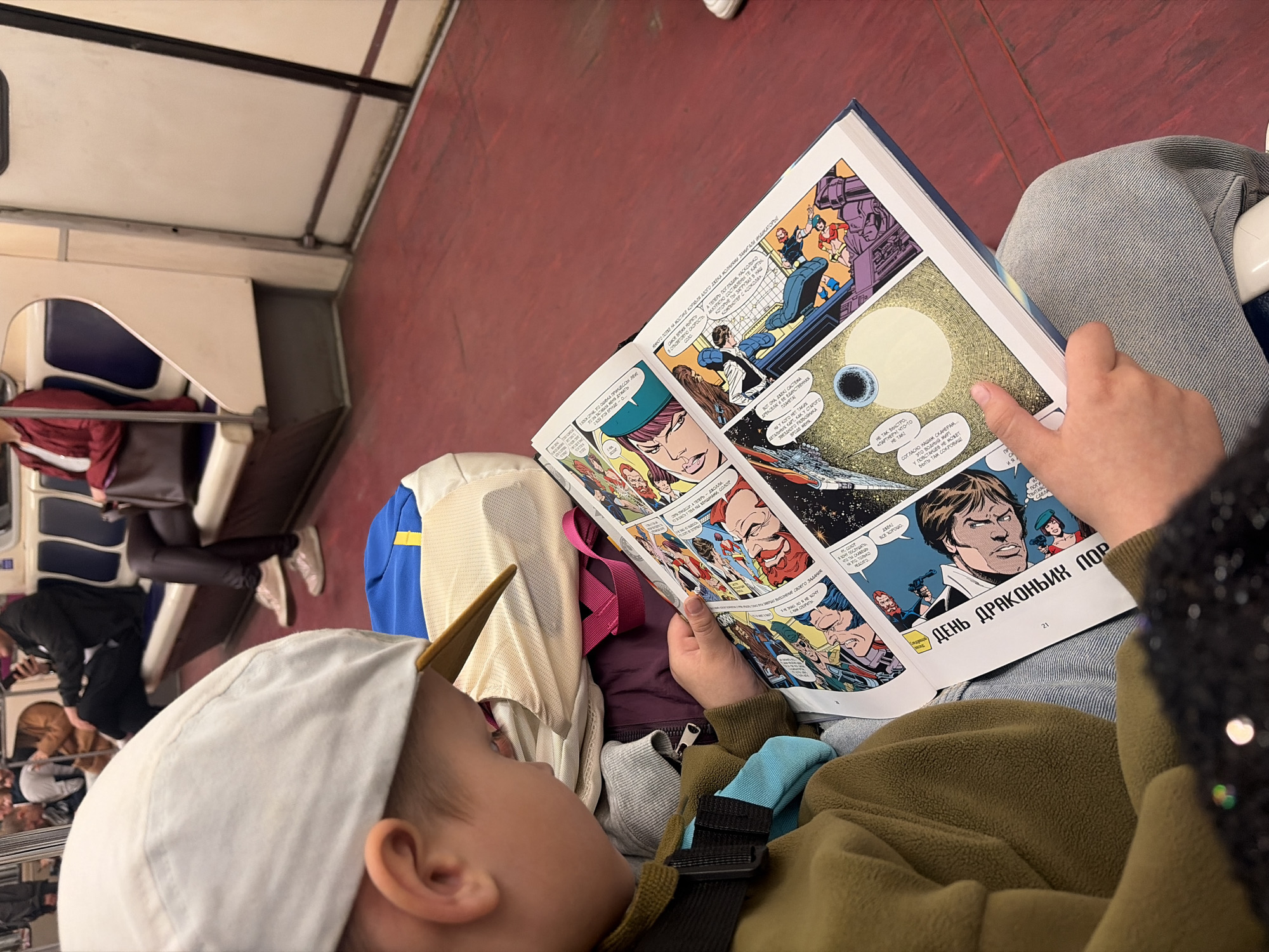 Here is a description of the image:
The image shows a child on what appears to be a train or subway car, possibly in St. Petersburg, Russia given the Cyrillic script in the book. The child, wearing a light-colored cap and an olive-green jacket, is engrossed in reading a comic book. The comic book is open, and shows panels with illustrations and text in Russian ("ДЕНЬ ДРАКОНЫХ ПОР" meaning "DAY OF DRAGON'S PORT"). The child's hand rests on the open page of the comic book.
Other people can be seen in the background sitting on seats of a subway car.