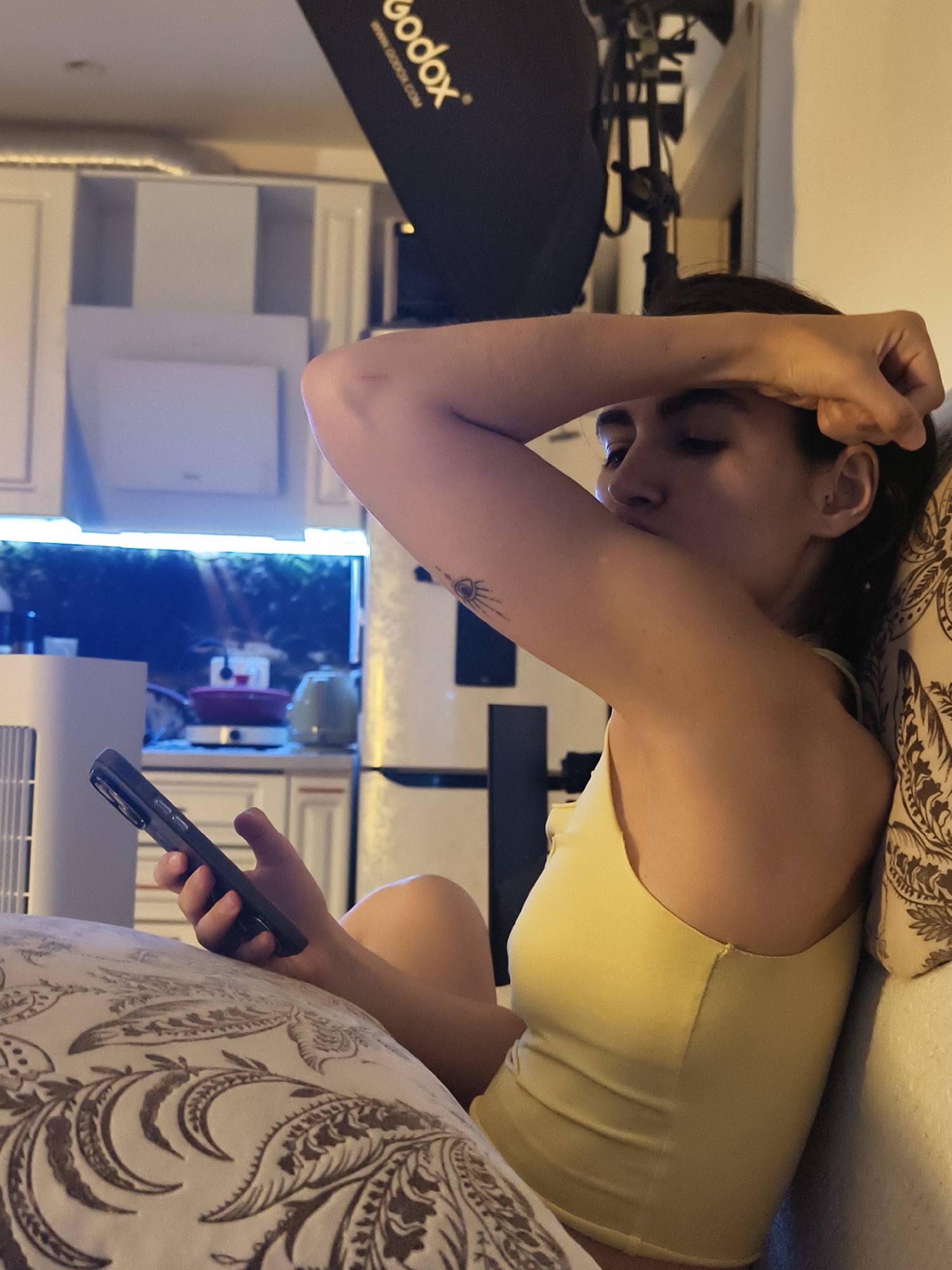 A female individual is depicted in a reclining position indoors, supported by patterned pillows. She wears a light yellow tank top. Her right arm is bent with the hand resting on her forehead, partially obscuring her face, while a small tattoo, resembling a stylized sun or eye, is visible on her right bicep. Her left hand holds a dark-colored smartphone, directed towards her gaze. The background reveals a kitchen area with white upper cabinets, under-cabinet lighting emitting a blue hue, and what appears to be an illuminated aquarium or terrarium behind the counter. On the counter, a red pot and a light green kettle are visible, with a white appliance partially on the left. A white refrigerator is positioned on the far right. Above the kitchen, a black softbox or studio light modifier bearing the "Godox" logo is present. The lighting is low, suggesting an evening or night setting. The image provides no visual information to determine the specific city or country, including Tyumen, Russia.