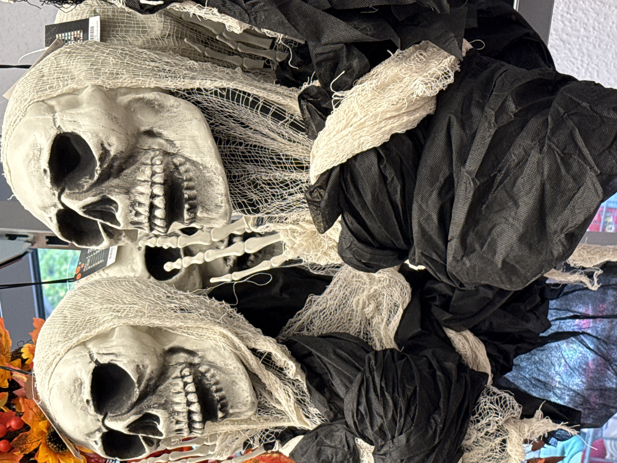 The image shows a close-up view of two Halloween decorations, specifically skeleton figures, in what appears to be a retail setting. The skeletons are made of white plastic, with detailed skull features and visible rib cages. They are draped in tattered white and black fabric, giving them a ghostly or grim reaper appearance. The foreground is dominated by the black fabric of the costumes, while the skeletons' heads and upper torsos are visible. In the background, some autumnal decorations with orange and yellow flowers and leaves can be seen, suggesting a seasonal display. The image is taken from a slightly low angle, focusing on the details of the decorations. The context of the city of Bad Lauterberg im Harz, Germany, is not visually present in the image.