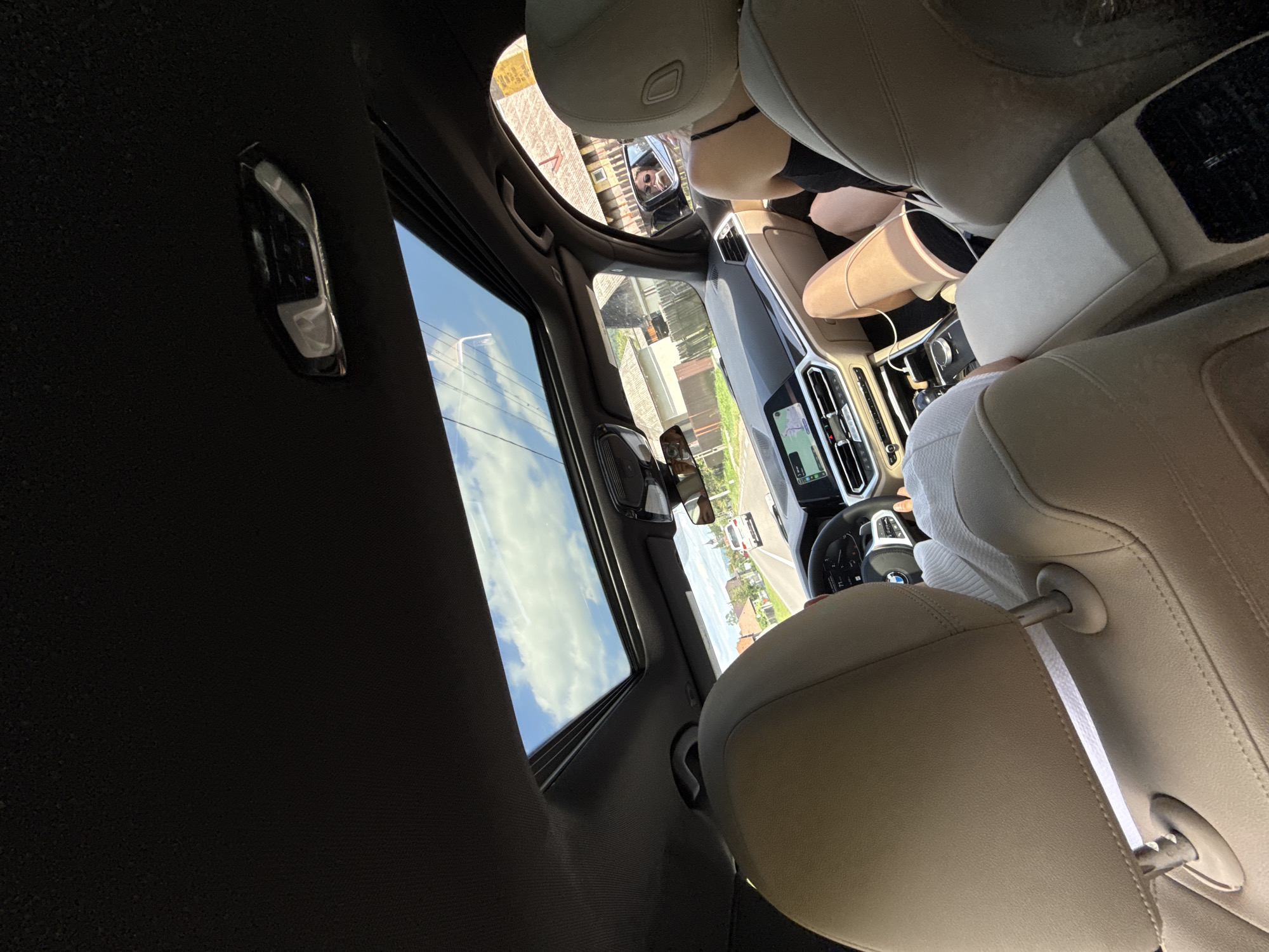 The image is taken from inside a car, looking forward through the open sunroof and windshield. The car's interior is visible, including the beige leather seats, dashboard with a navigation screen, and a steering wheel. Two individuals are in the car; one is driving and another, a woman wearing a black top and shorts, is seated in the passenger seat. The woman's legs are crossed and illuminated by sunlight. Through the sunroof, a bright blue sky with white clouds is visible, and the car is driving on a road in Hârja, Romania, with houses and trees lining the street. The road appears to be a rural or suburban area.