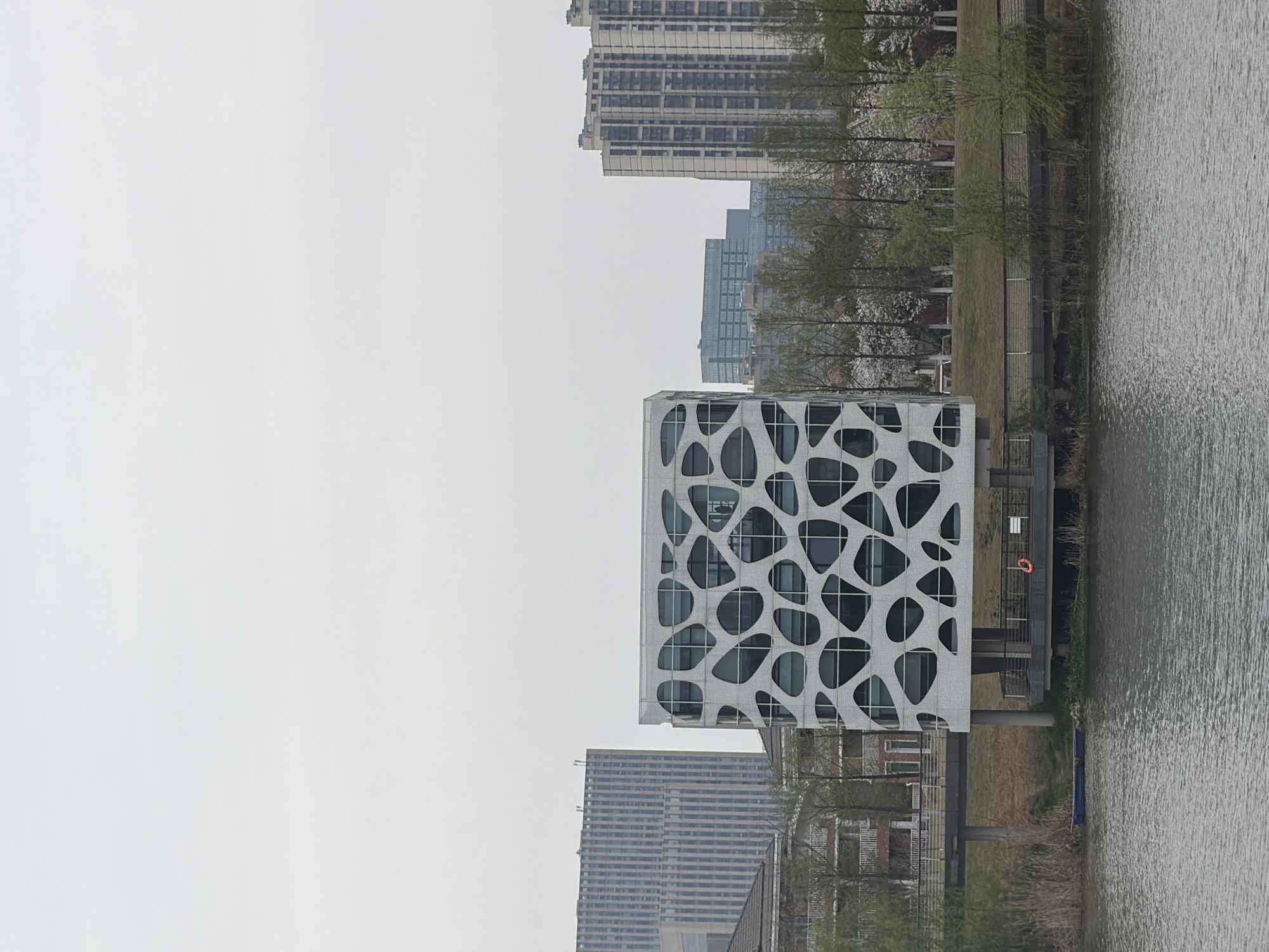 The scene portrays an urban waterfront in Jiangning District, China, under an overcast sky. A prominent multi-story building with a distinctive white facade, characterized by numerous irregular, organic-shaped cutouts revealing dark-tinted windows, stands elevated on stilts above a concrete pier. An orange life buoy is secured to a black railing on this pier. To the left of the main building, a small blue boat is partially visible at the water's edge, partially obscured by reeds. A pedestrian bridge extends horizontally behind the modern building. In the background, a tall, vertically-striped building is visible to the left, alongside a lower residential structure with balconies. To the right, a line of trees, some bearing white blossoms, forms a park-like boundary along the water, with several distant high-rise buildings in the far background. The foreground consists of dark, rippled water. No individuals, specific actions, or interactions are discernible.