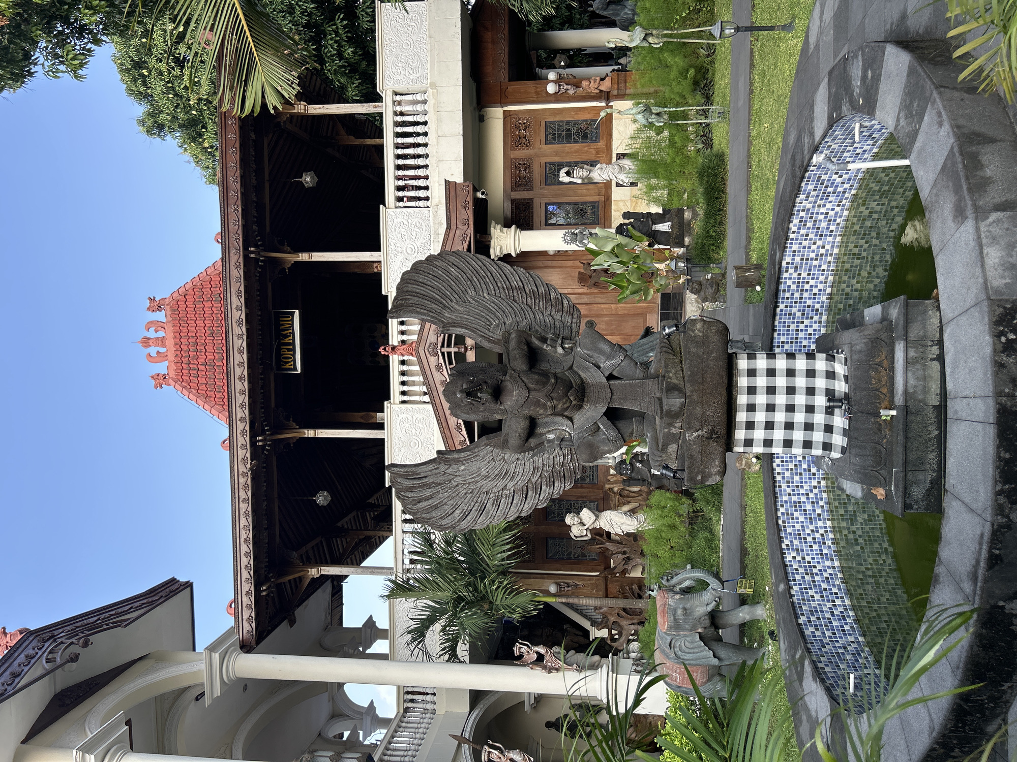 This is a medium shot of the exterior of a building that appears to be a cafe, possibly called "Kopi Kamu" judging by the sign. The building has a traditional architectural style, featuring a red tiled roof with decorative elements, wooden beams, and an upper balcony with white railings. The foreground is dominated by a circular fountain with blue and white tiled walls. A stone statue of a mythical winged creature, possibly Garuda, stands in front of the fountain, draped with a black and white checkered cloth. To the left of the fountain is a stone statue of an elephant. The background shows lush green vegetation and trees. The lighting suggests it is either early morning or late afternoon. The location is identified as Yogyakarta, Indonesia, suggesting a cultural or tourist-oriented establishment.