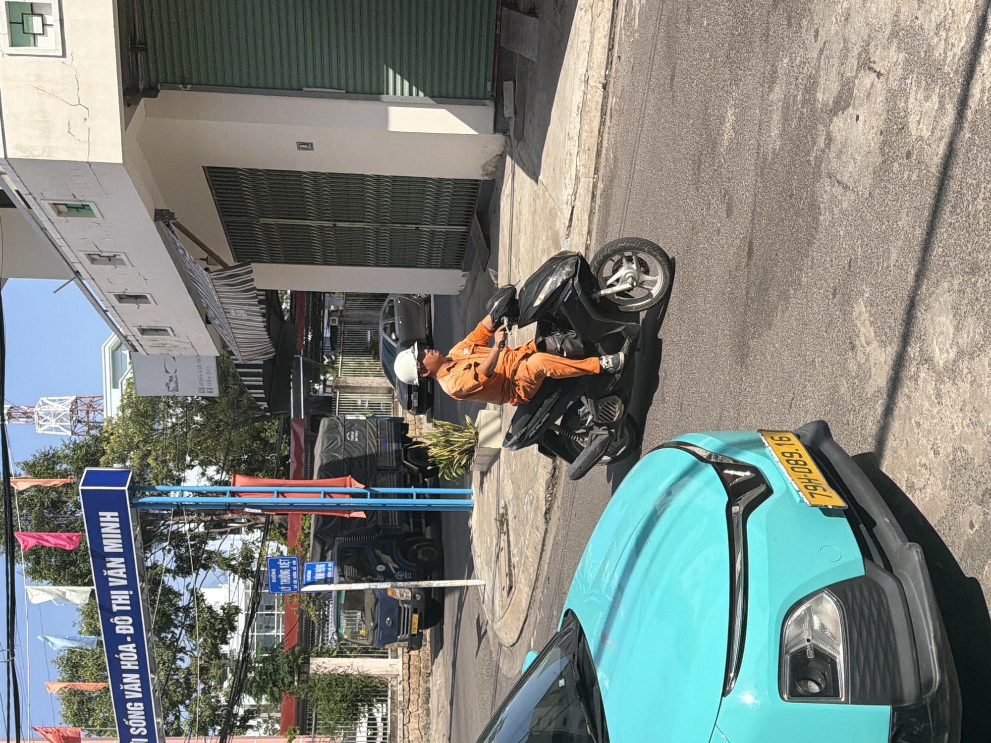 In the city of Nha Trang, Vietnam, on a sunny day, a man wearing an orange uniform and a white helmet is riding a black motorcycle. He is on a paved road, with a turquoise car in the foreground to his left. Behind him, a blue truck is parked, and further back, a building with green metal shutters is visible. A street sign with Vietnamese text is also present in the scene. The environment suggests an urban setting with typical street elements.