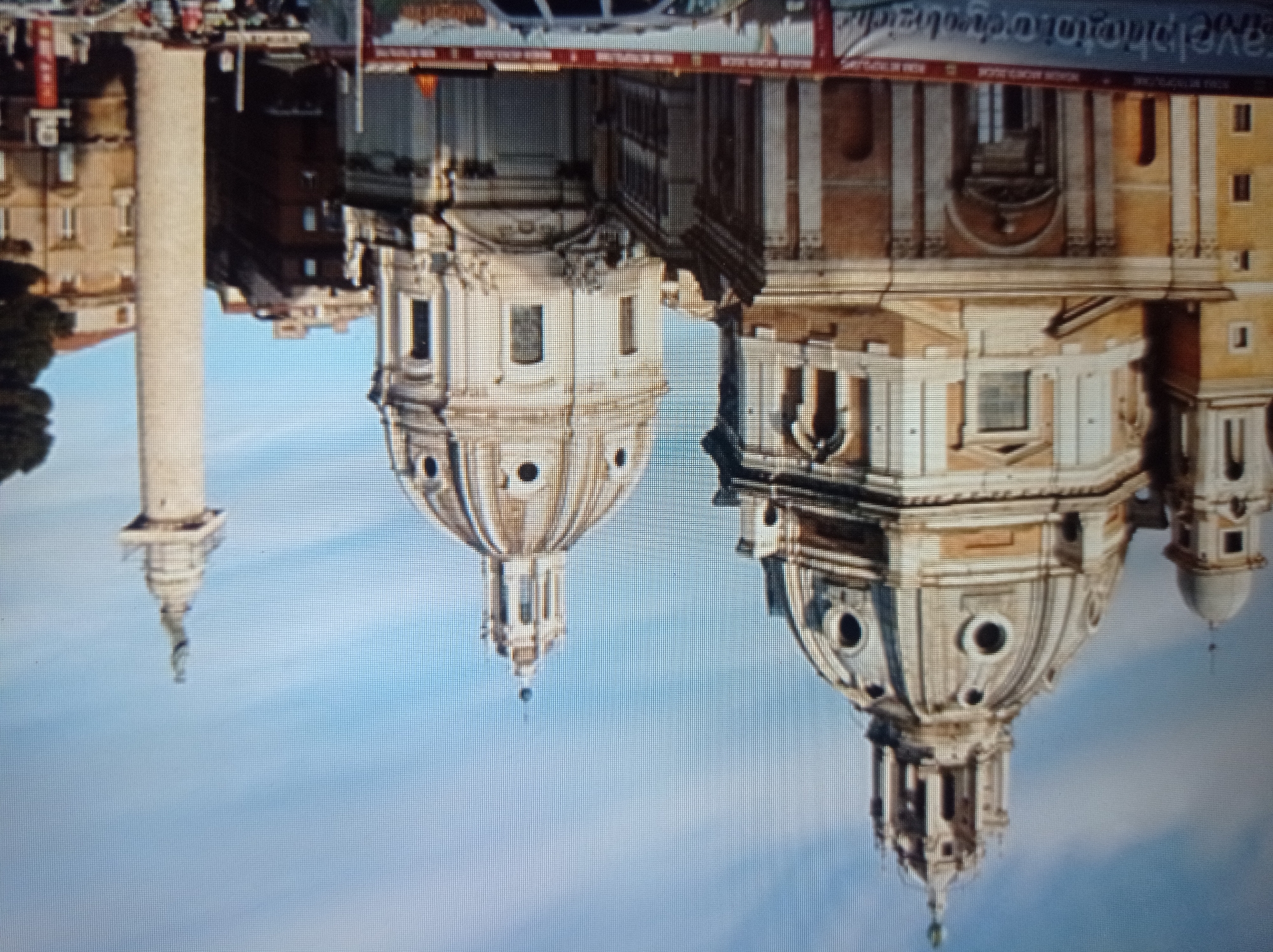 The image shows a wide shot of two large domed buildings and a tall column with a statue on top, set against a blue sky with some clouds. The buildings are ornate, with classical architectural features, including domes, columns, and windows. The column appears to be made of stone and is a prominent feature on the right side of the image. In the foreground, there is a banner with text that is partially visible, and some people and vehicles are present, suggesting a public space. The overall scene depicts a historical or cultural landmark, likely in a European city. The city of Mukachevo, Ukraine, is not depicted in this image.