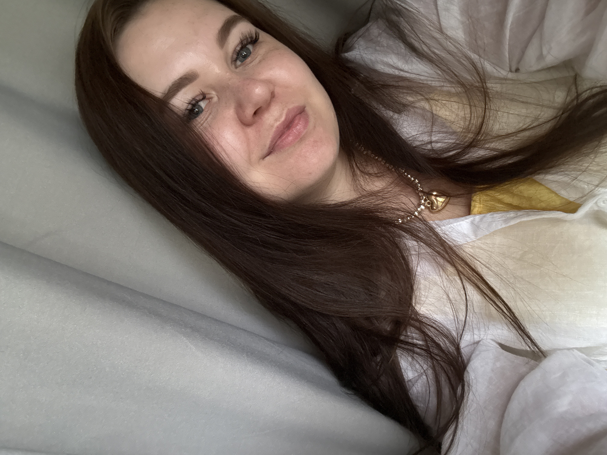 A young woman with long, dark brown hair and blue eyes is lying down, looking at the camera. She is wearing a white shirt and a yellow garment underneath, and a gold necklace with a heart-shaped pendant. The background appears to be curtains or draped fabric. The scene is likely indoors, possibly a bedroom, and no specific location in Chaikovsky, Russia can be identified from the image.