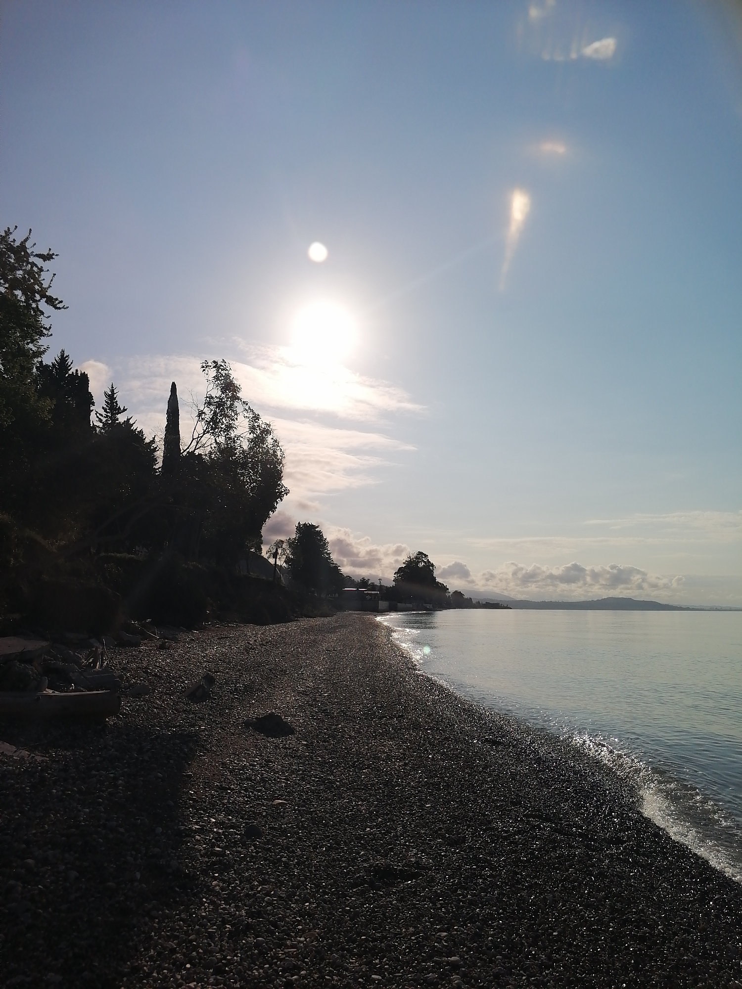 The image captures a serene coastal landscape during what appears to be early morning or late afternoon, characterized by strong backlighting from a low sun.
The scene features a wide pebble beach in the foreground, stretching from the lower left to the middle distance on the right. The pebbles vary in size and are dark, reflecting the light from the sun, particularly where the gentle waves lap the shore. To the left, a densely vegetated hillside rises, covered with dark, silhouetted trees, including several tall, slender coniferous or cypress-like trees and other deciduous foliage. A small, light-colored boat or object is partially visible on the beach near the base of the hillside on the far left.
The calm, clear water of a sea or large lake occupies the right side of the image and extends into the background. Small, white-capped waves are visible only at the immediate shoreline. In the mid-ground, across the water, low-lying hills or mountains are faintly visible under a hazy sky.
The sky is bright and clear blue, with scattered, wispy clouds. The sun is a dominant feature, positioned high in the mid-left portion of the sky, creating a strong lens flare and washing out part of the upper right sky. The intense sunlight backlights the entire scene, casting the hillside and most of the beach into deep shadow, while the sun's reflection glimmers on the water's surface.
There are no people or discernible activities taking place. No visible text is present in the image. The overall impression is one of peaceful natural beauty with dramatic lighting.
