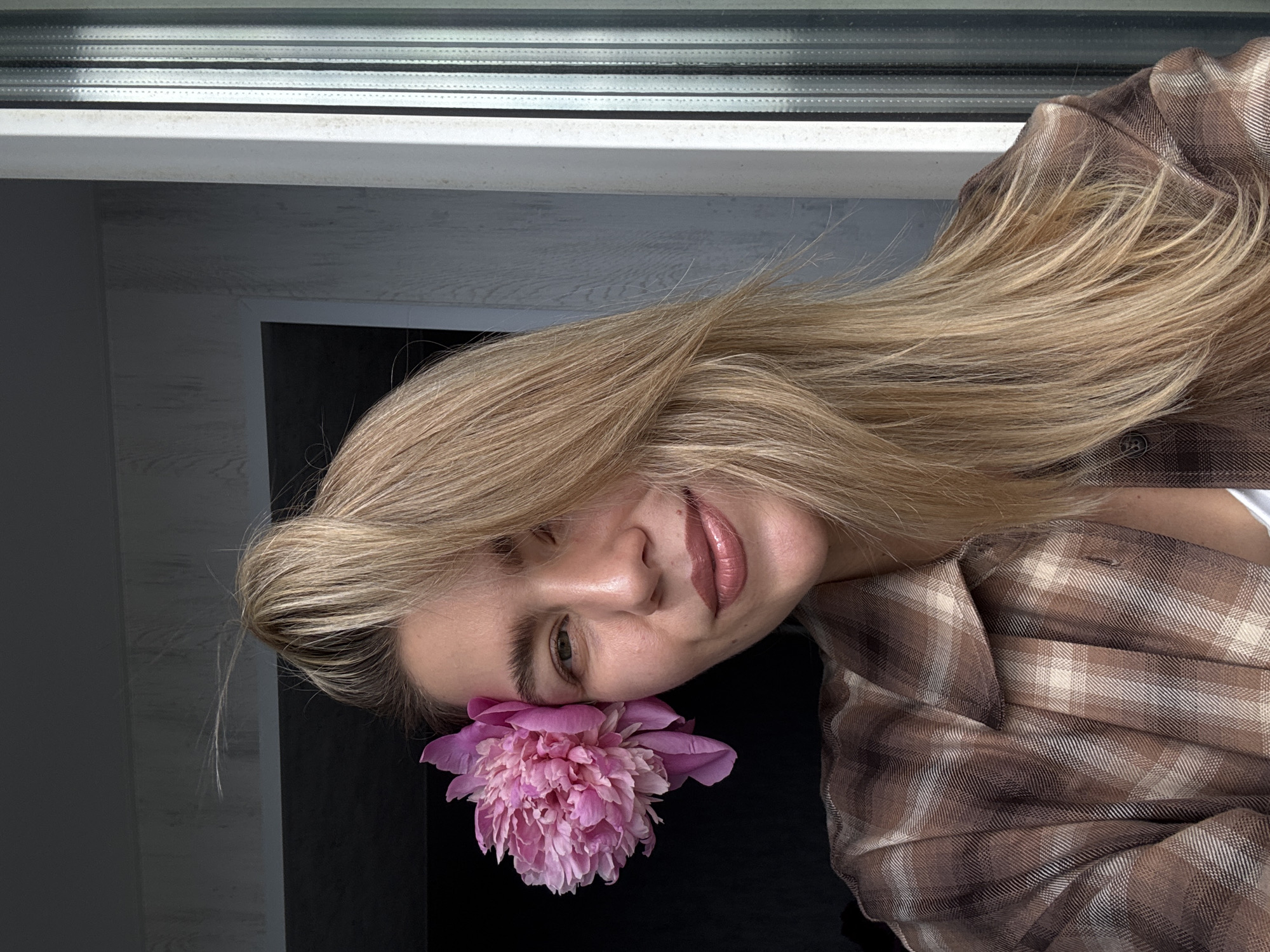 A woman with long, light blonde hair, partially tied into a high bun with loose strands framing her face, looks directly at the camera with a gentle smile. She wears a brown and tan plaid button-down shirt, with a white undershirt visible below. A large, multi-petaled pink peony flower is tucked behind her left ear. Her lips appear glossy, and her visible left eye is green. The background to her immediate left is dark and indistinct. To her right, a light-colored, possibly textured wall is visible, along with a white window frame and a pane featuring vertical textured patterns. The scene is illuminated by natural light, seemingly from the right side of the frame, indoors, in the city of Unknown, Unknown.