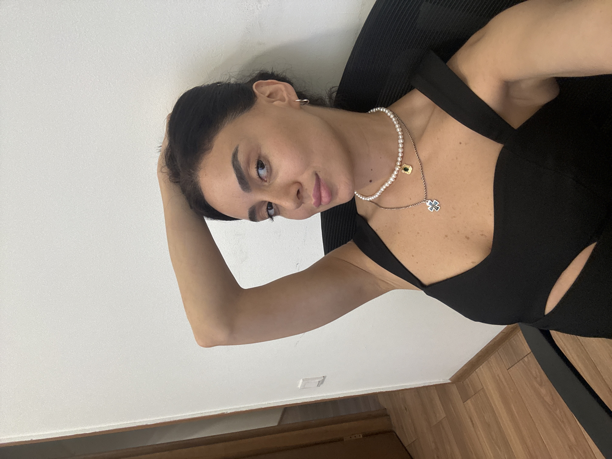 A woman is sitting in a chair indoors, with her left arm raised and her hand resting on the back of her head. She is wearing a black dress and two necklaces, one of pearls and a pendant of a green gem, and another with a cross pendant. She is looking at the camera with a slight smile. The background is a white wall and a wooden door and floor are visible on the left side of the image. There is no indication of the location being Tbilisi, Georgia.