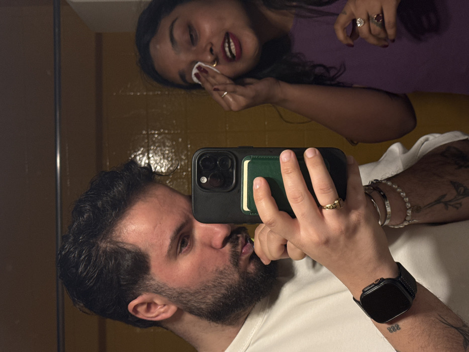 A selfie captures two people in a mirror. A man with a beard, wearing a white shirt and a smart watch on his left wrist, holds the phone, obscuring part of his face. Above him, a woman with dark hair wearing a purple shirt cleans her face with a cotton pad. Gold and silver jewelry adorns both individuals. The background shows a yellow-tiled wall. The selfie captures a casual, intimate moment.