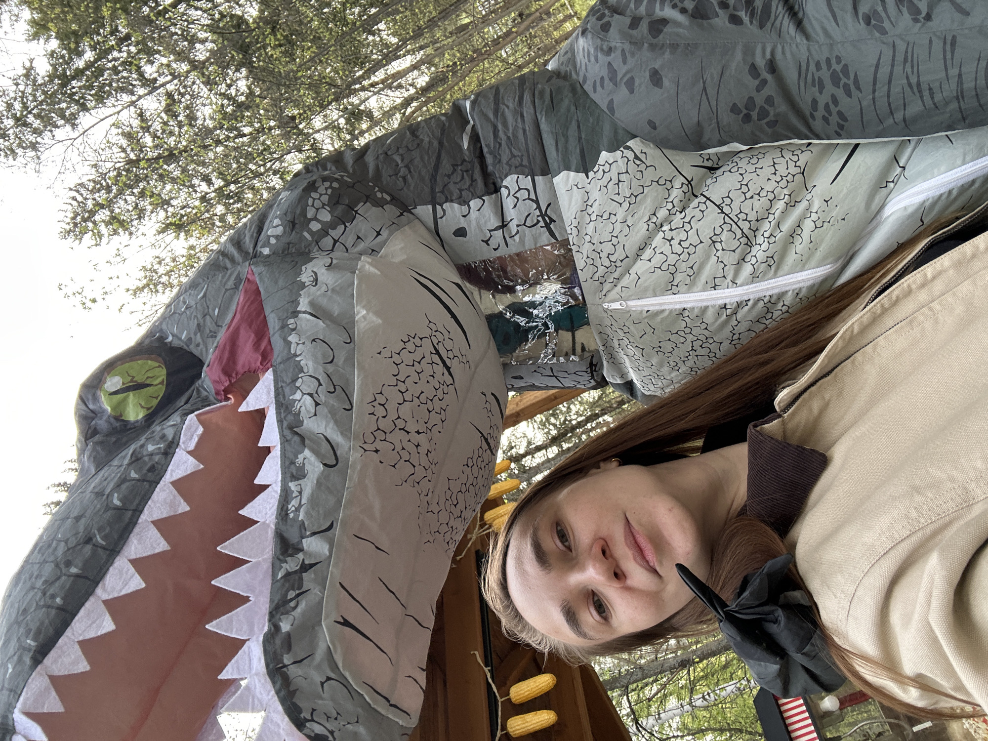A person is posing with a large, inflatable dinosaur costume outdoors. The costume is gray and white with a menacing open mouth displaying sharp, triangular white teeth. The person is wearing a tan jacket with a dark collar. Behind the dinosaur costume, trees and the sky are visible. A wooden structure with dried corn cobs hanging from it can be seen in the background. The photo appears to be taken from a low angle. The location is not identifiable as Izhevsk, Russia based on visual cues.