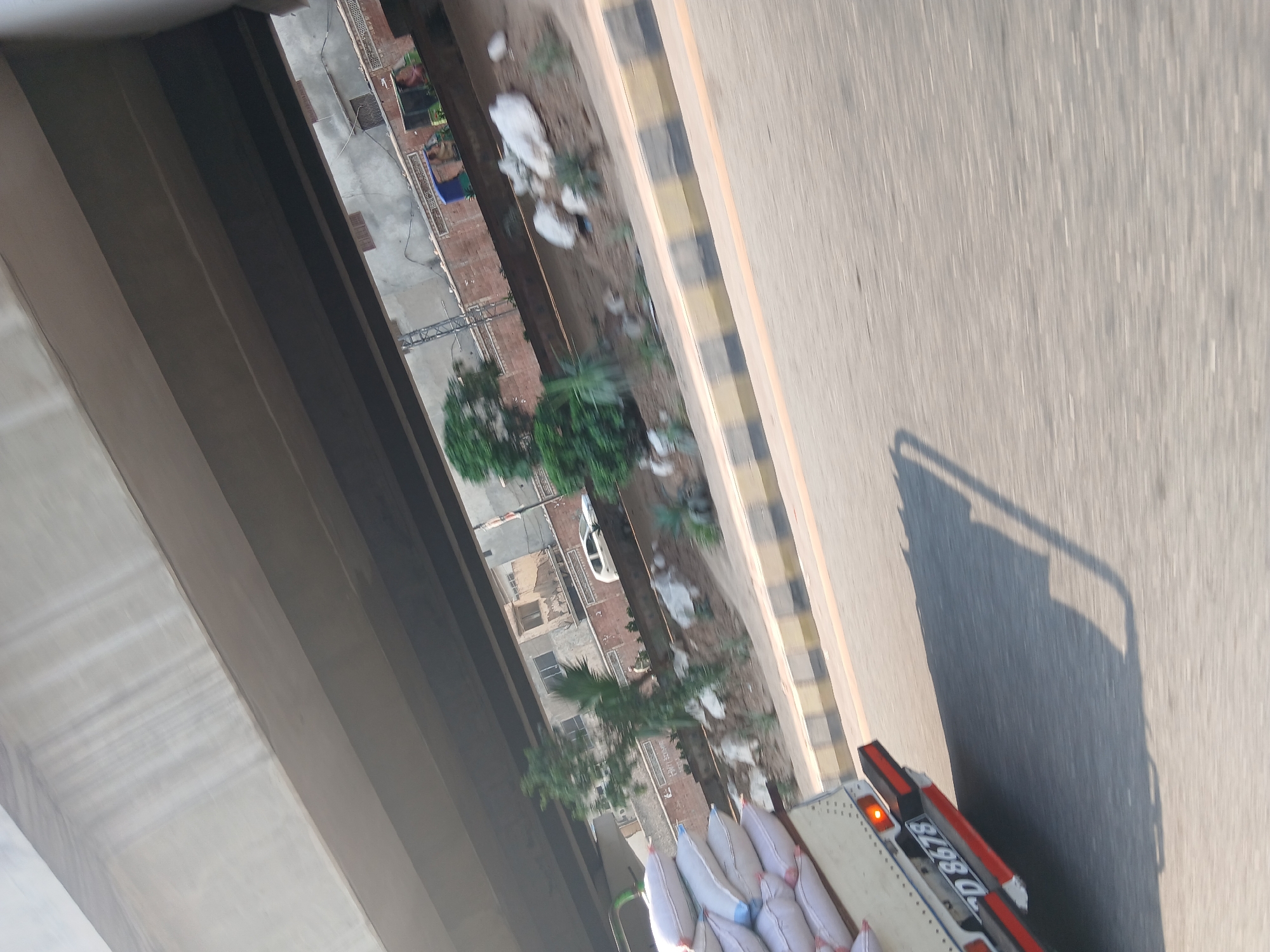 A slightly blurred, eye-level shot captured from a vehicle in Peshawar City Tehsil, Pakistan, shows a road with traffic and infrastructure. In the foreground on the left, the back of a white truck is visible, loaded with numerous white sacks. The truck has a license plate reading "D 8678". The road surface is asphalt, with a yellow and black striped median barrier.
Above the road, a large concrete overpass casts a shadow. Behind the median, a slope leads up to a row of buildings constructed with reddish-brown bricks and concrete. Several trees and bushes are planted on the slope, with some larger plants and rocks scattered around. A white car is visible on a street behind the median. Further in the background, a blue cart is present with two individuals. The overall scene suggests a typical urban environment with ongoing traffic and construction.