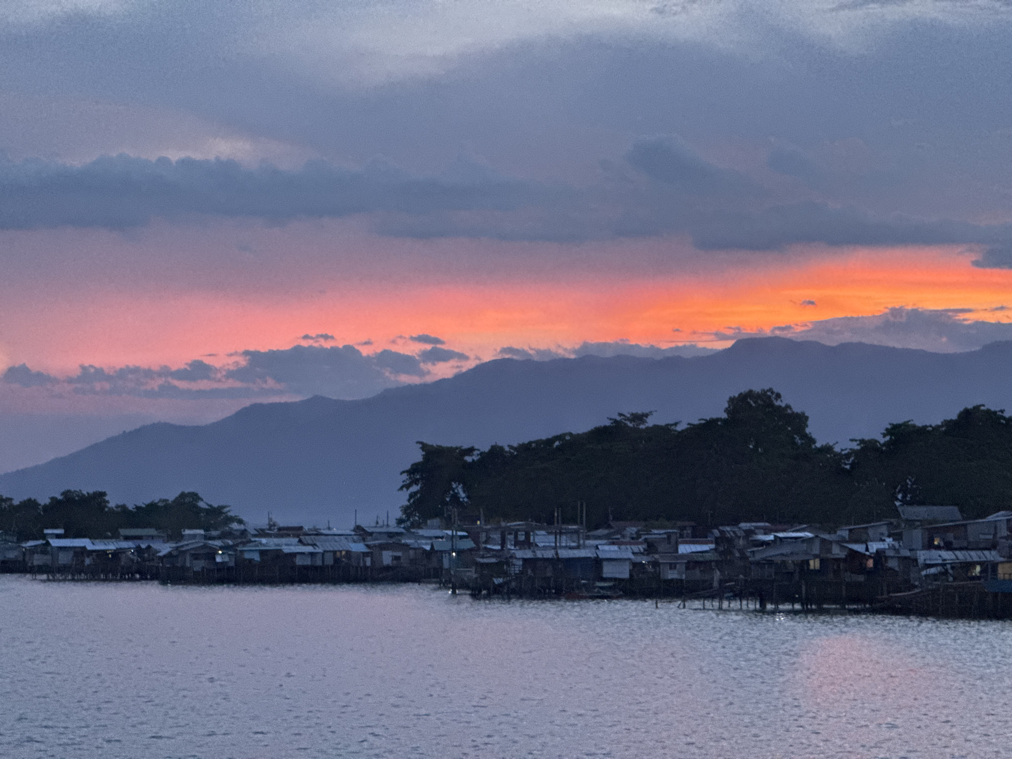 The image shows a sunset over a body of water in Davao City, Philippines. In the foreground, there are numerous shacks built on stilts over the water. Behind the shacks, there is a line of trees, and further back are silhouetted mountains. The sky transitions from orange and red near the horizon to gray and purple above. The water is a light blue-gray color. There are no people visible in the image. The overall scene suggests a coastal slum area.