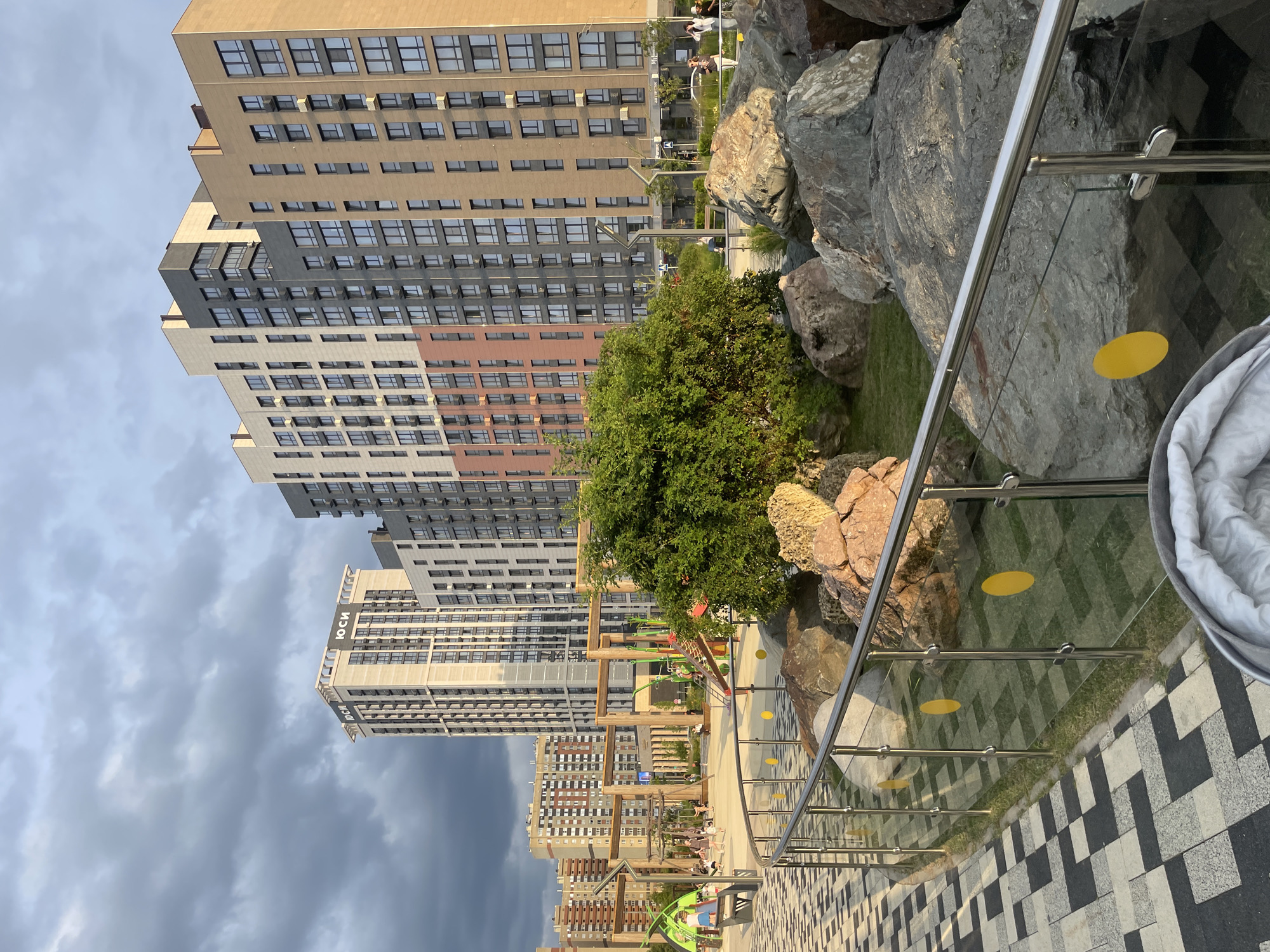 The image shows a modern residential complex in Stavropol, Russia. In the foreground, a paved walkway with a black and white checkered pattern leads towards a playground area. A metal and glass fence separates the walkway from a landscaped area with large rocks and green grass. A large green bush is prominent in the mid-ground. Behind the playground, several tall residential buildings with diverse architectural styles and colors dominate the skyline. The sky is partly cloudy with dramatic shadows. Several people are visible in the playground area, some appearing to be children playing. The overall scene depicts a contemporary urban environment with amenities for residents.