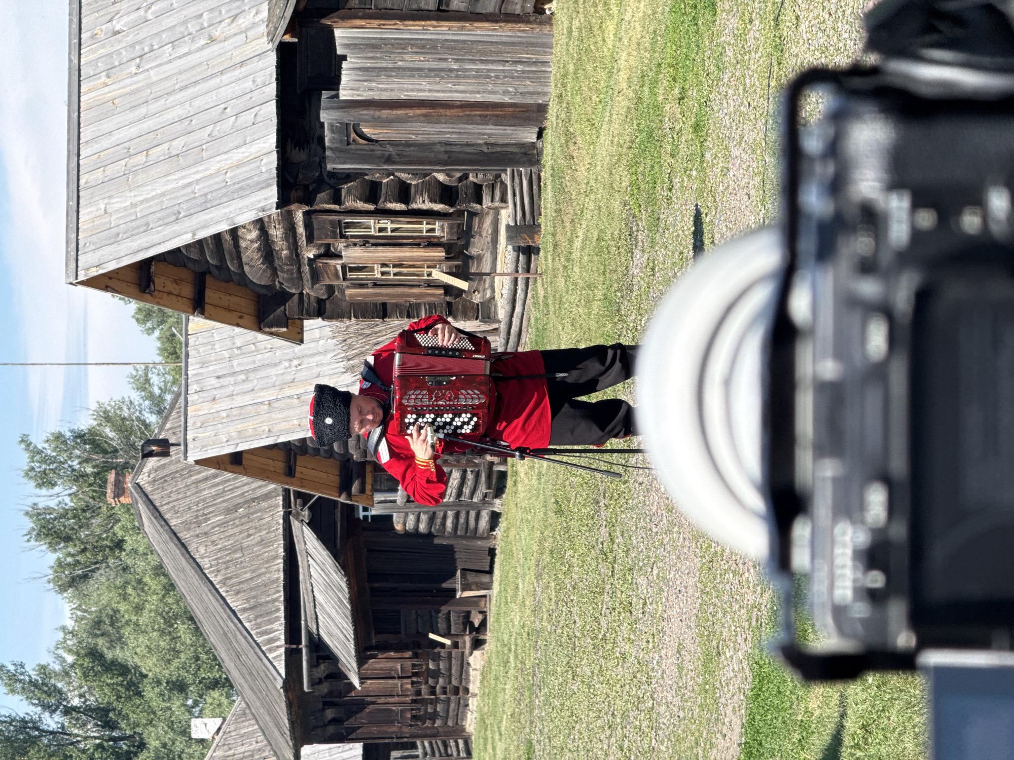 In the city of Shushenskoye, Russia, a man dressed in a traditional red Cossack outfit with a black fur hat is playing an accordion. He is standing in front of several old wooden log houses with weathered wooden roofs. The houses appear to be part of a historical village or museum. The ground is covered in green grass and gravel. A camera lens is visible in the foreground, out of focus, suggesting the scene is being filmed or photographed. The sky is clear and blue with scattered clouds, and trees are visible in the background. The man is holding the accordion with both hands and appears to be actively playing it.