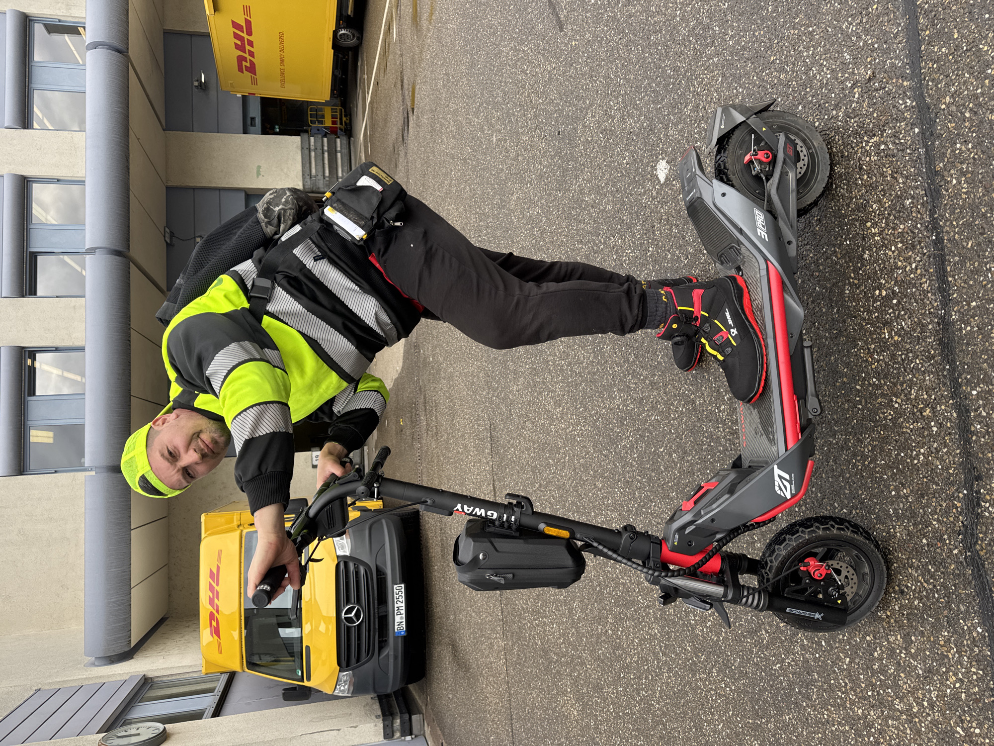 An outdoor, eye-level shot, rotated to be upright, shows a man posing with a black, red, and grey electric scooter on asphalt pavement. The man, positioned centrally, is smiling directly at the camera. He wears a high-visibility neon yellow and black jacket with reflective stripes, black trousers, black and red sneakers, and a yellow cap. He stands with his left foot on the scooter's deck, holding the handlebars.
The electric scooter features "TIGWAY" on its stem, "3 PRO" on the main body, and "XSEGRIDE" on the front fender. It has large, rugged tires with disc brakes and red calipers. A small black storage bag is attached to the scooter's stem.
In the background, two yellow DHL vehicles are visible. One is a large DHL truck with "DHL" and "EXCELLENCE. SIMPLY DELIVERED." emblazoned on its side. The other is a DHL delivery van, identifiable as a Mercedes-Benz Sprinter by its grille and logo, with the German license plate "BN PM 2550". A modern building with grey vertical columns and windows stands behind the vehicles.
The scene is set on damp asphalt, suggesting overcast weather or recent rain, during daytime. This logistics or industrial area is located in Bietigheim-Bissingen, Germany.