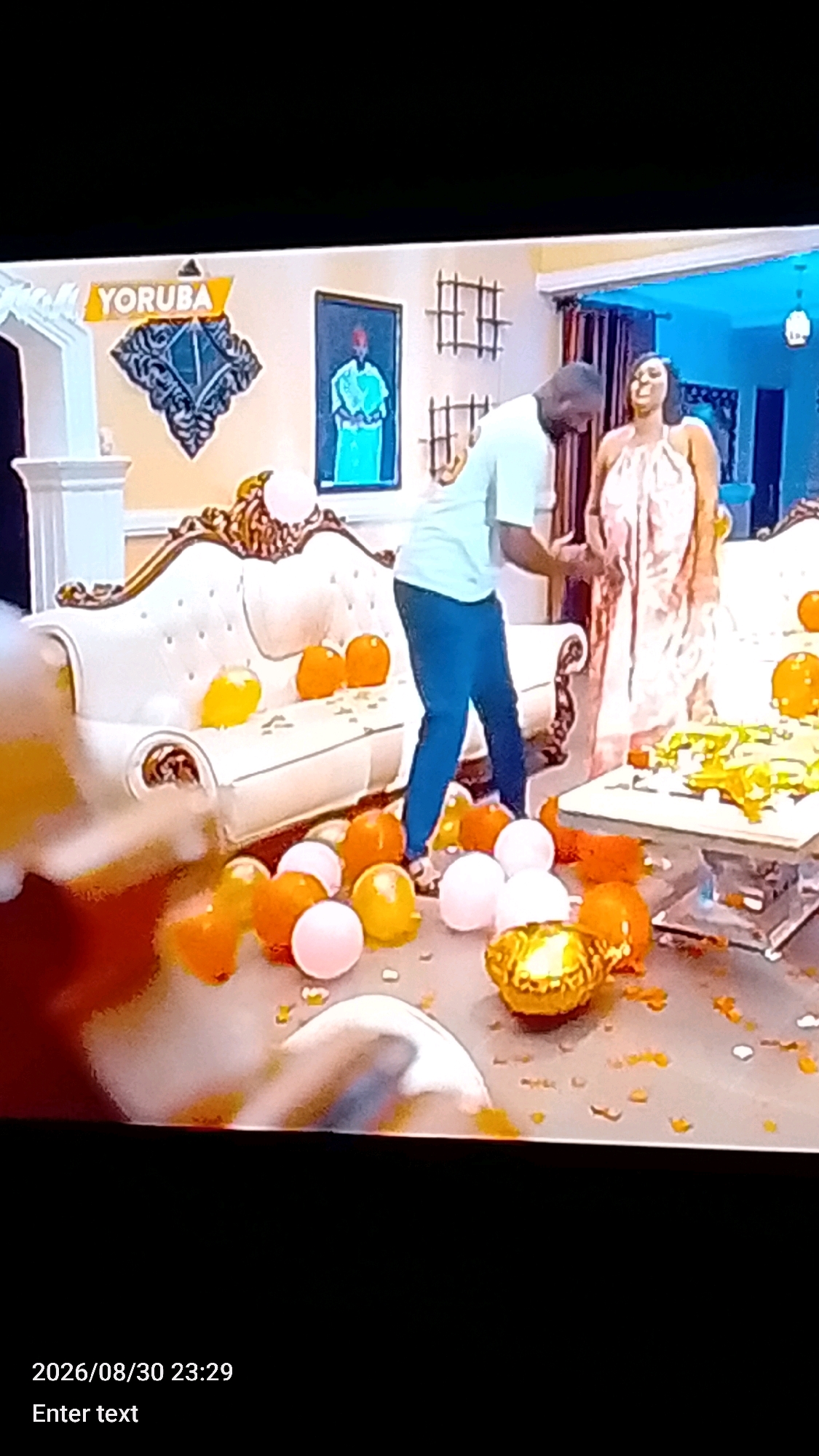 The image depicts an indoor scene, possibly a living room, decorated with numerous balloons, primarily in orange, pink, and gold. A white, ornate sofa is visible in the foreground, with some of the balloons placed on it and scattered around it. In the background, two people are present. One person, a man in a light-colored t-shirt and blue pants, is leaning over, possibly interacting with the balloons. The other person, a woman in a light-colored, flowing dress, is standing and looking towards the man, with her mouth open as if laughing. A framed picture of a person in traditional attire hangs on the wall, along with decorative elements resembling window panes. A low coffee table in the mid-ground also has balloons and decorative confetti on it. The overall ambiance suggests a celebratory or party setting. The timestamp at the bottom indicates "2026/08/30 23:29." The mention of "Lokoja, Nigeria" in the prompt is not visually confirmed by any element within the image itself.