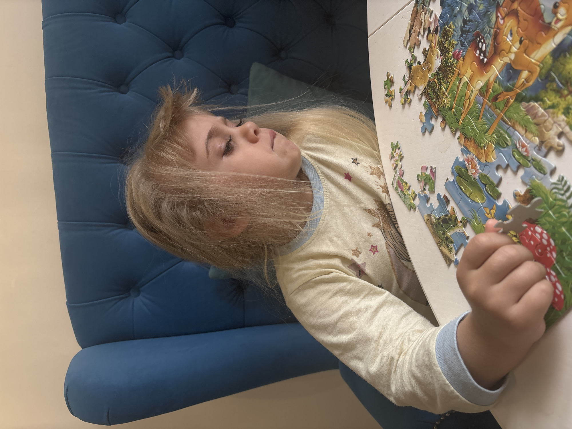 A young girl with long, blonde hair is lying on a blue, tufted couch, engaging with a partially completed jigsaw puzzle. The puzzle features an image of Bambi and other woodland elements. The girl is wearing a light-colored long-sleeved shirt with star patterns. She is holding a puzzle piece in her right hand. The scene is set indoors against a light-colored wall. The specific city and country are unknown.