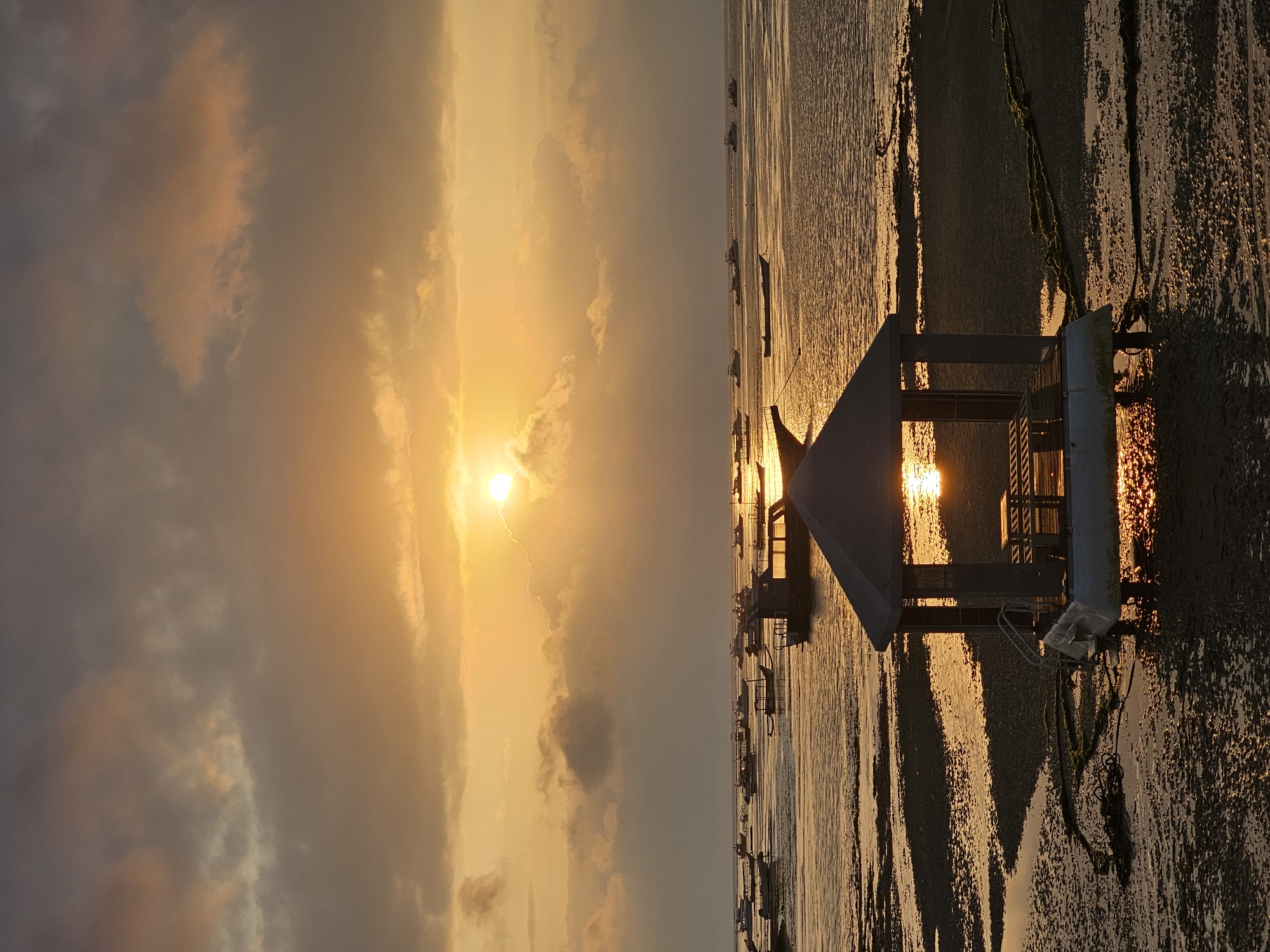 This is a vertical, medium shot of a sunset over a body of water with boats docked near a structure. The sun is a bright yellow orb in the upper center of the frame, partially obscured by clouds. Rays of sunlight emanate from the sun, illuminating the clouds and the water with a golden hue. In the foreground, a small, octagonal gazebo with a peaked roof sits on a raised platform in the shallow water. The gazebo has open sides and a wooden table and benches inside. A set of stairs leads down from the platform into the water. The water is calm and reflects the golden light of the sunset. The seabed is visible in places, with sand and patches of seaweed. Numerous boats of various sizes are moored in the water in the middle ground and background. They are mostly silhouetted against the bright sky. The scene appears to be a coastal area, likely a harbor or bay. Given the context of Denpasar, Indonesia, this scene could be located in a popular tourist area with boat services, such as near Sanur beach or a similar coastal location within the city. The overall mood of the image is peaceful and serene.