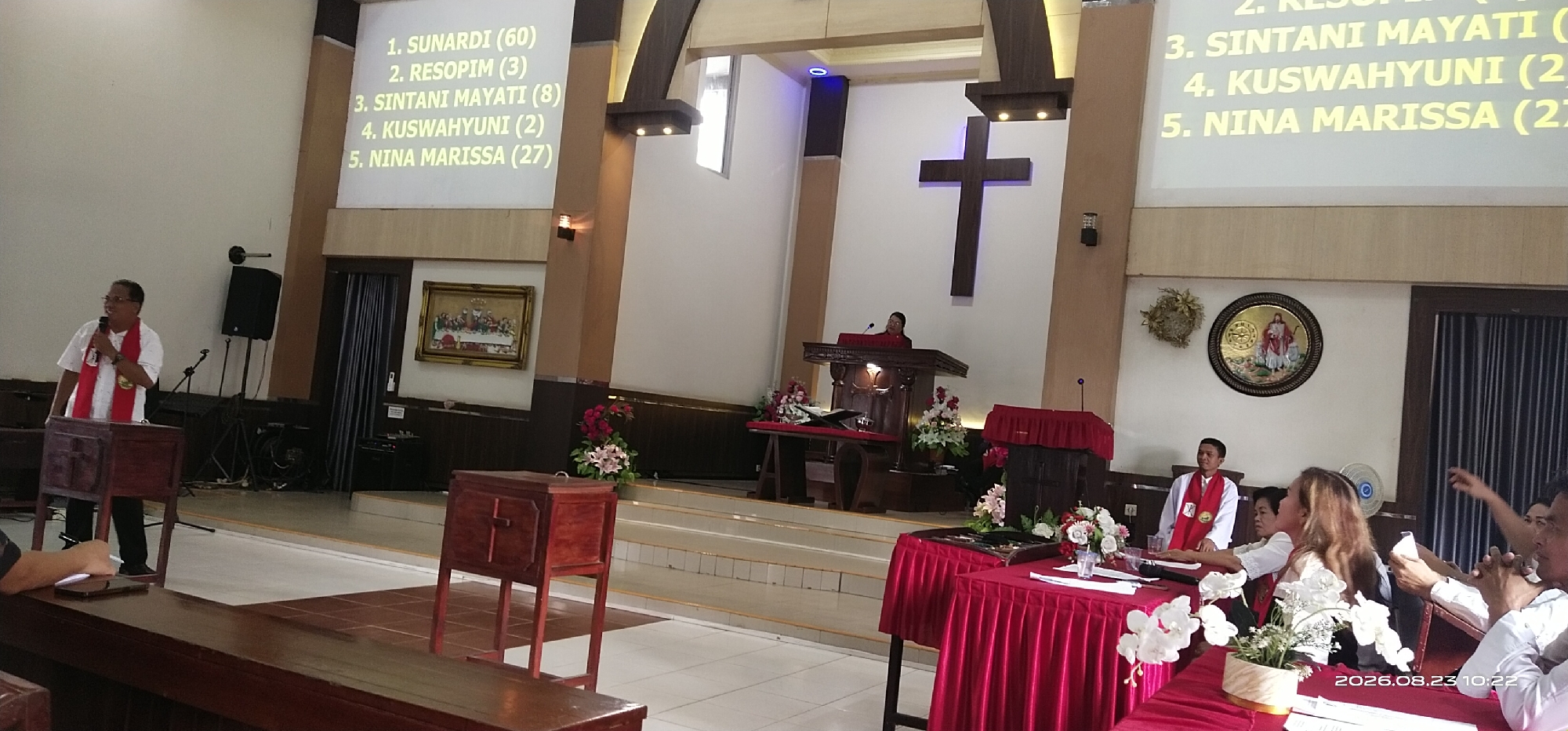 The image depicts an indoor gathering, likely a religious service, within a church setting. A projection screen displays a numbered list of names with numbers in parentheses, suggesting results or rankings. Several individuals are present, with one man in a white shirt and red stole speaking into a microphone at a wooden podium, positioned on a raised platform. Another person, also wearing a red stole, is seated at a table covered with a red tablecloth. The environment features wooden pews, a large cross mounted on the wall, and floral arrangements. The date stamp at the bottom right indicates August 23, 2026, at 10:22 AM. The location is identified as Jaar, Indonesia.