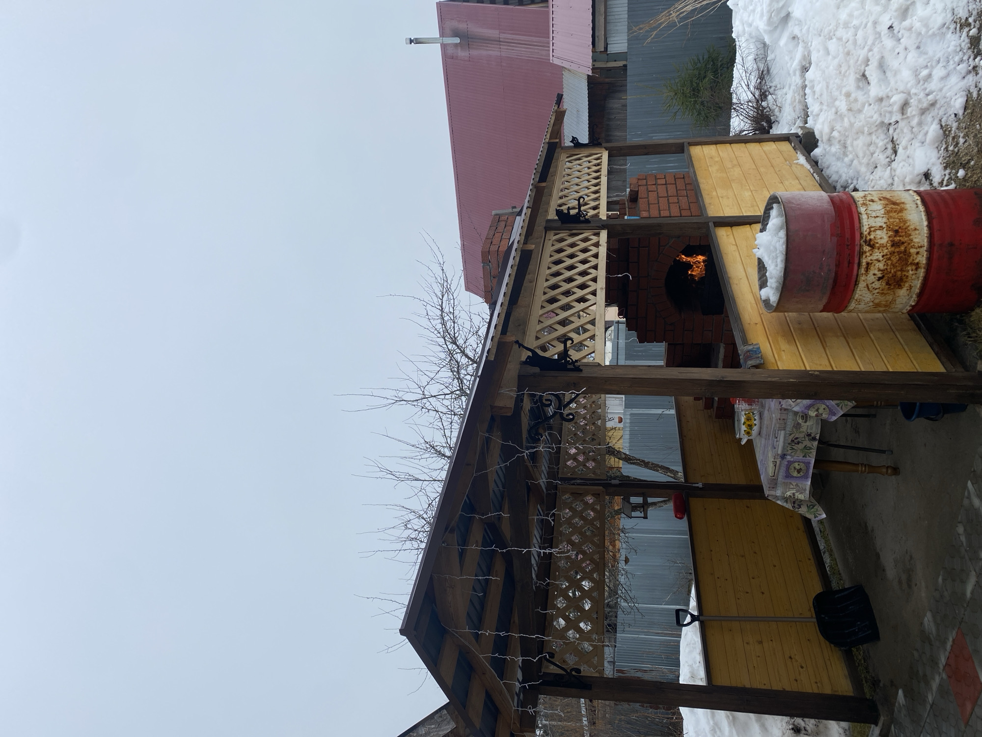 The image shows an outdoor gazebo structure in Chaikovsky, Russia. The gazebo has a wooden frame with latticework panels and decorative metal hooks along the roofline. Bare tree branches are visible near the roof. A brick barbecue grill with visible flames is within the gazebo. A red and white barrel is positioned beside the grill, partially filled with snow. Outside the gazebo, there is snow on the ground. A building with a red roof is visible in the background. Inside the gazebo, there is a table covered with a patterned tablecloth. A black snow shovel is leaning against the side of the gazebo.