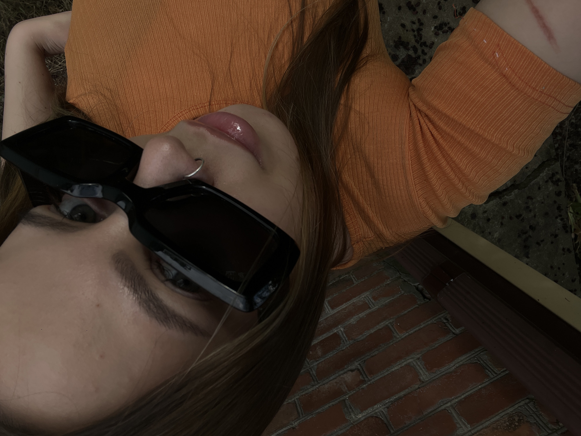 The image shows a close-up, high-angle shot of a young woman wearing an orange ribbed t-shirt and black sunglasses. She has a nose ring and appears to be looking at the camera, with her lips slightly parted. Her left forearm is visible, showing a red mark that appears to be a scar. The background consists of a brick wall and what looks like a downspout. The lower part of the image shows a patchy ground with some dry vegetation. The woman's hair is brown and falls over her right shoulder. The image does not contain any information that can be used to determine the location in the city of Novopokrovskaya, Russia.