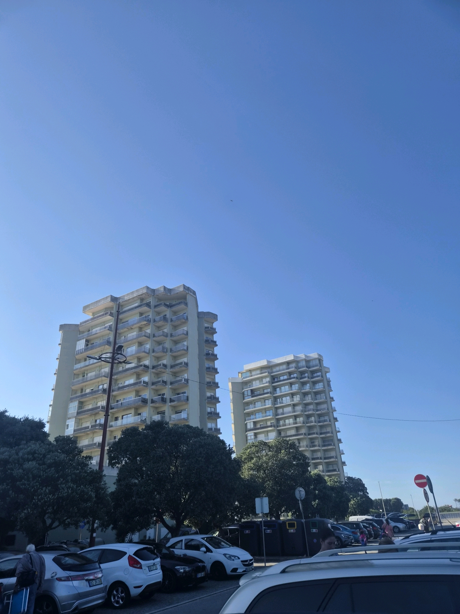 The image depicts two modern apartment buildings with balconies, set against a clear blue sky. In the foreground, a parking lot is visible with several cars, including a silver hatchback and a white hatchback, parked. Trees with dark green foliage are situated between the parking lot and the buildings. A person with a backpack and luggage is standing near the silver car. Traffic signs are also present, including a red and white circular sign with a horizontal bar, indicating prohibition. The scene appears to be in the city of Esposende, Portugal.