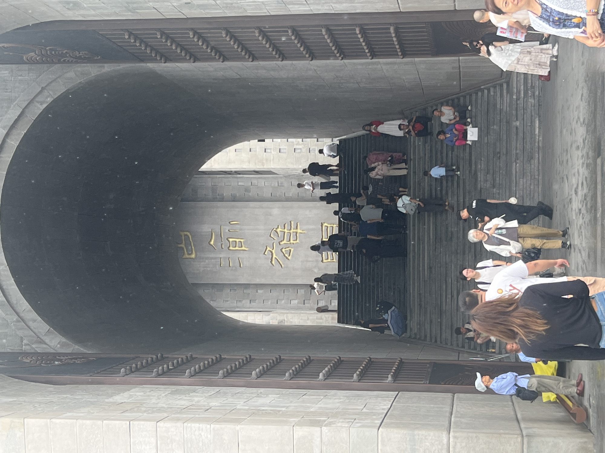 The image shows a wide stone staircase leading up to a grand, arched entrance. The entrance is flanked by large, ornate stone pillars with intricate carvings and vertical decorative elements. Above the archway, large golden Chinese characters are inscribed on a stone facade. Numerous people are present, ascending and descending the stairs, or standing and sitting on them. Some individuals are looking towards the camera, while others are engaged in conversation or looking at their surroundings. A few people are wearing casual clothing, while others appear to be dressed more formally. The overall scene suggests a public gathering or a tourist attraction. The location is Yuzhong District, China.