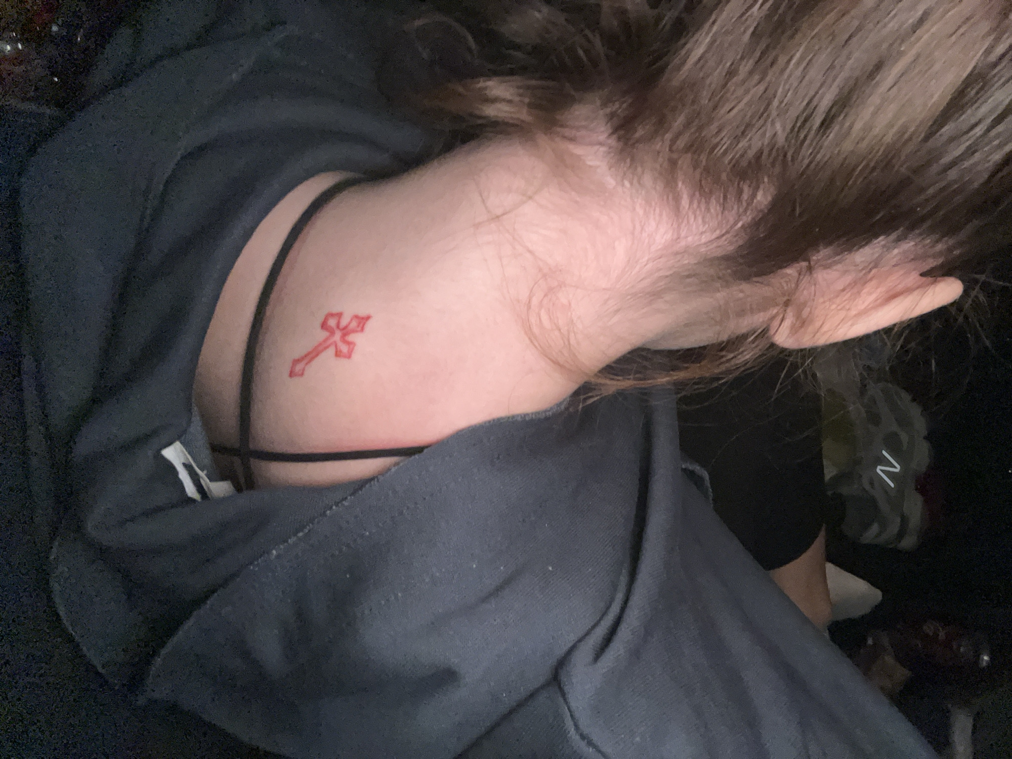 A close-up, overhead shot shows a person lying down wearing a dark grey hooded sweatshirt. Visible skin includes their neck, the top of their chest, and the side of their face and ear. A thin black strap of clothing (likely a bra or tank top) is visible on their chest. A small red cross tattoo is located on the side of their neck, near the collarbone. The person has brown hair. A leg covered in black fabric and a white shoe are visible in the background on the right side of the image. There is no information in the image to suggest the location is Varna, Bulgaria.