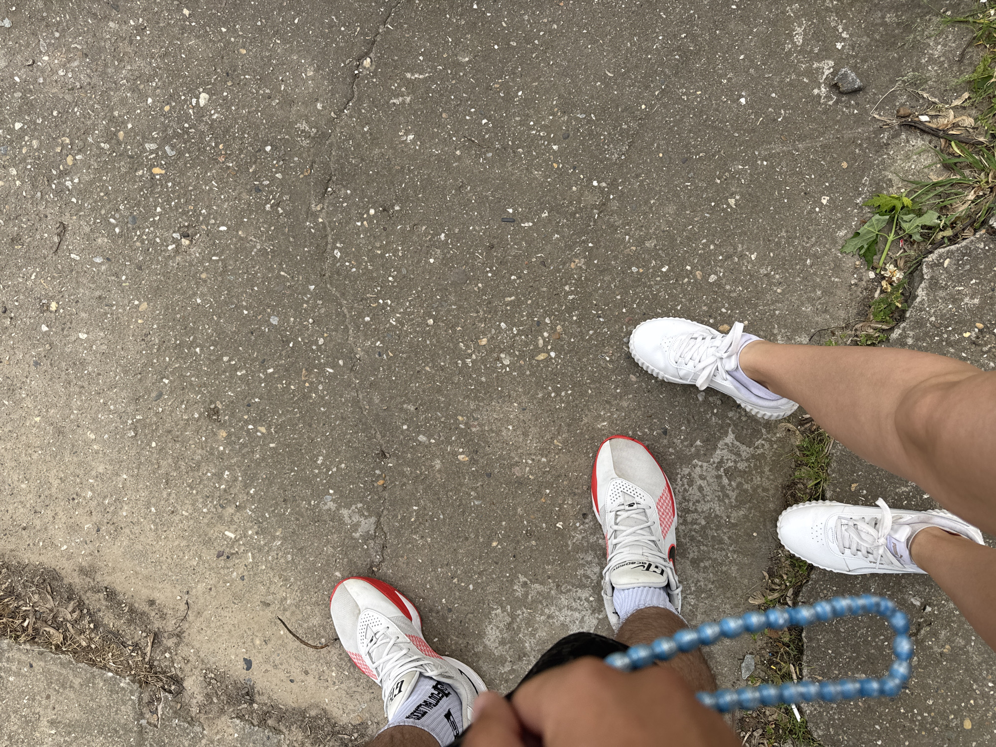 Two pairs of feet are positioned on a concrete surface. The concrete shows wear and tear with cracks and a small patch of green growth near the top right. One person is wearing white and red athletic shoes with white socks, holding a string of blue beads. Another person is wearing white shoes. The location appears to be Glojene, Bulgaria.