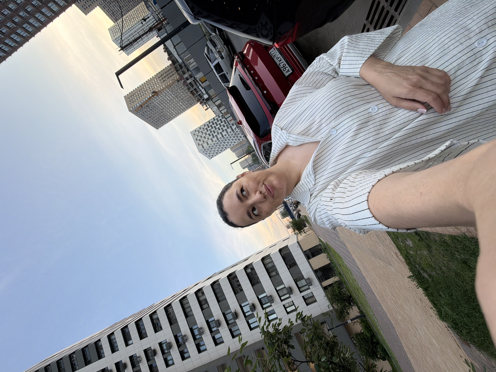 A woman with dark hair pulled back poses for a selfie outdoors, facing the camera. She wears a white shirt with thin black pinstripes and buttons. Her left arm is extended, holding the camera. In the background, several modern, tall residential buildings with numerous windows are visible, some under construction. Red and black cars are parked on the street, and a paved walkway runs through a grassy area. The sky is clear with a hint of sunset hues. The scene appears to be in a contemporary urban residential development in Moscow, Russia, given the architectural style of the buildings and the presence of Russian license plates on the vehicles.