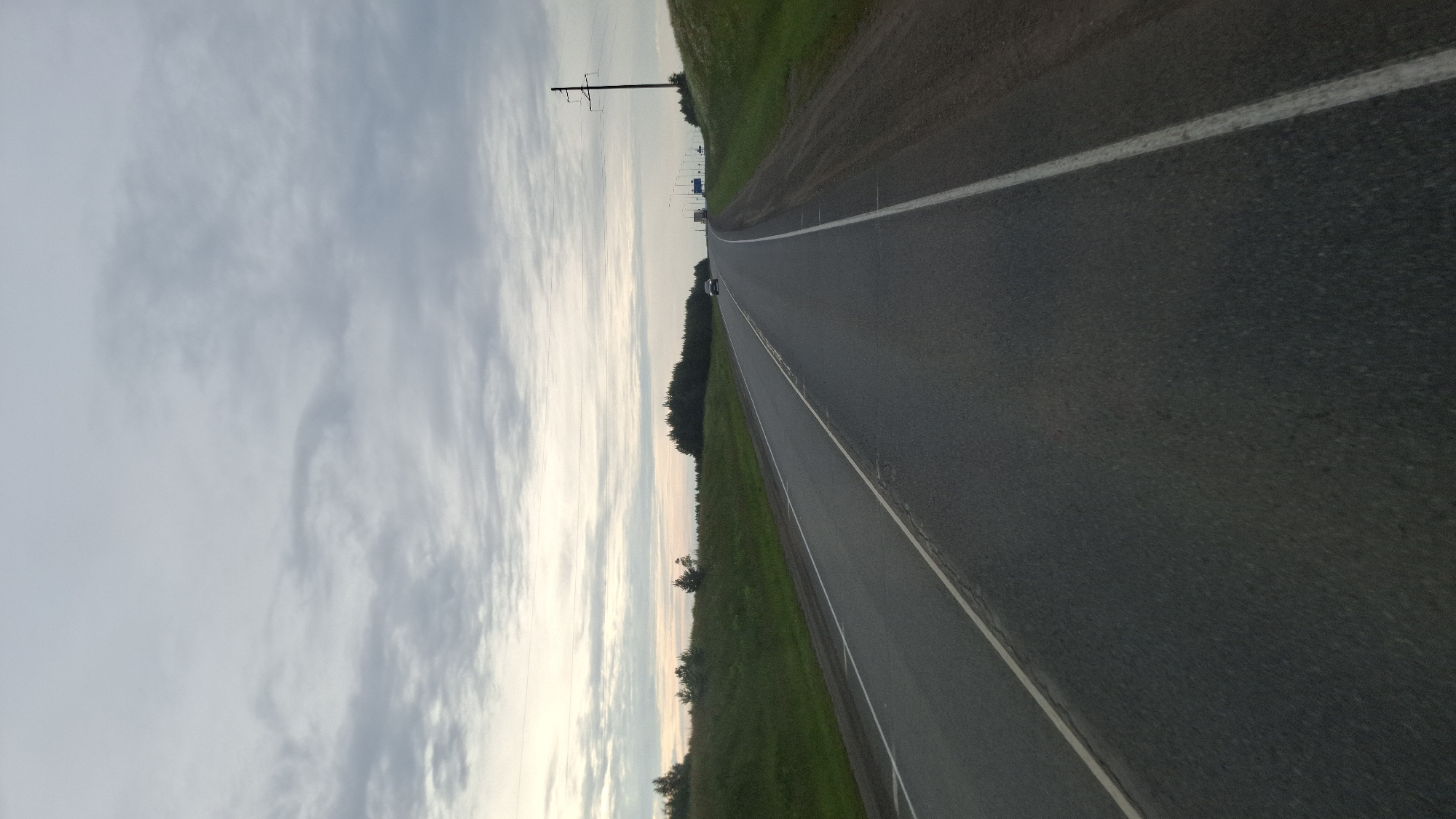 The media file depicts a horizontal, wide-angle view of a road stretching into the distance. The road is paved and has a white line marking its center. To the right of the road is a grassy embankment. To the left of the road, there is a line of trees and then a vast, cloudy sky. The sky is a mix of light grays and whites, suggesting it might be overcast or nearing dusk. A solitary white car is visible on the road in the distance. Power lines and a utility pole are also present along the left side of the road, near the embankment. The overall scene is a quiet, rural landscape, possibly in Berezovo, Russia, given the location context. The lighting suggests it is either early morning or late evening. There is no visible text in the image. The mood is serene and somewhat empty.