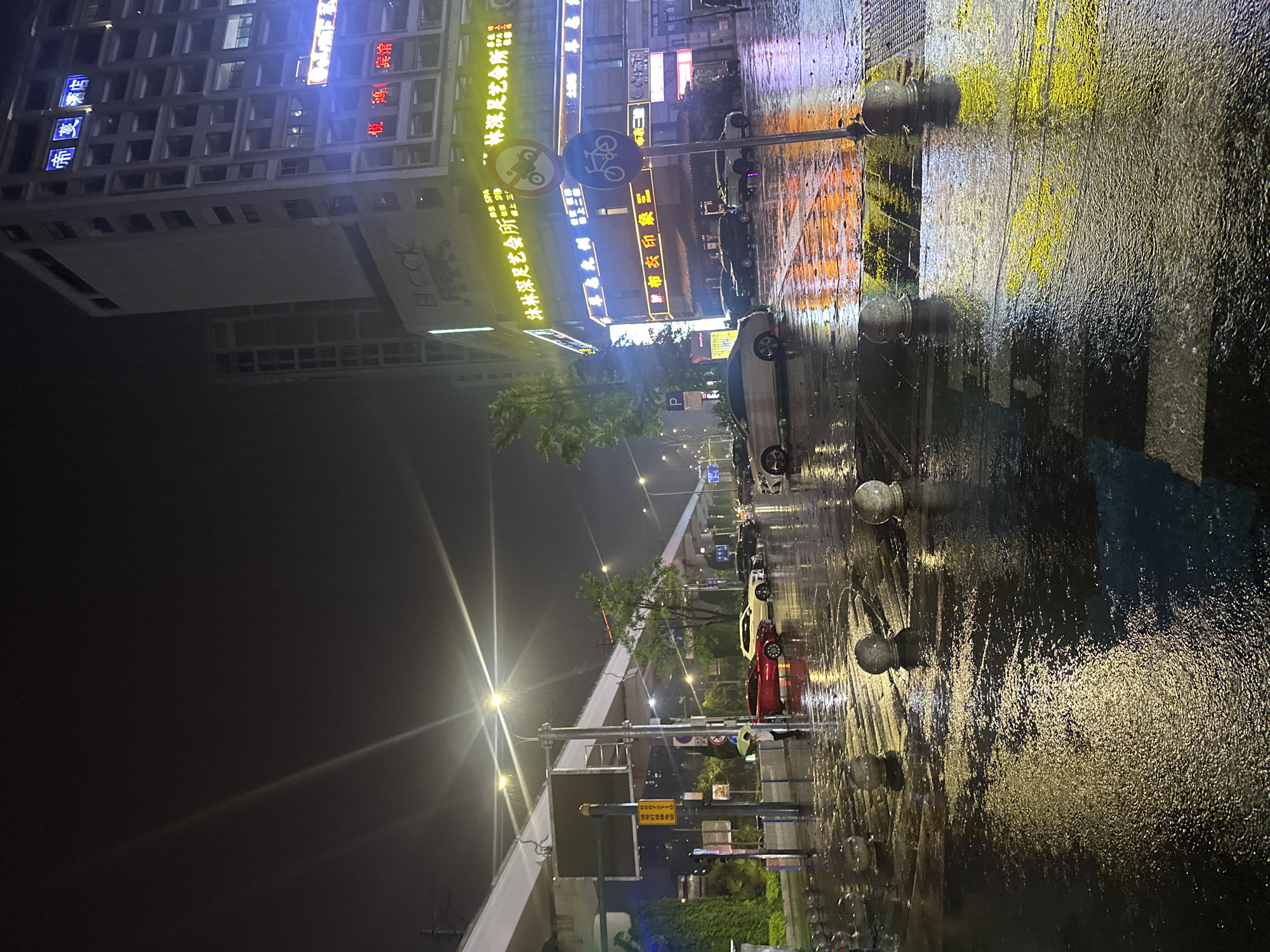 The image depicts a nighttime urban street scene in Chongqing High-tech Zone, China, after a rain. The road is wet, reflecting the lights from nearby buildings. Several cars are parked along the street. Buildings with illuminated signs in Chinese characters and English letters line one side of the street. A bicycle lane sign is visible. Pedestrian walkways are lined with decorative bollards. Street lights illuminate the scene, and a pedestrian overpass is visible in the distance. A sign shows the telephone number, 68072110 for traffic signal repair, along with Chinese text.