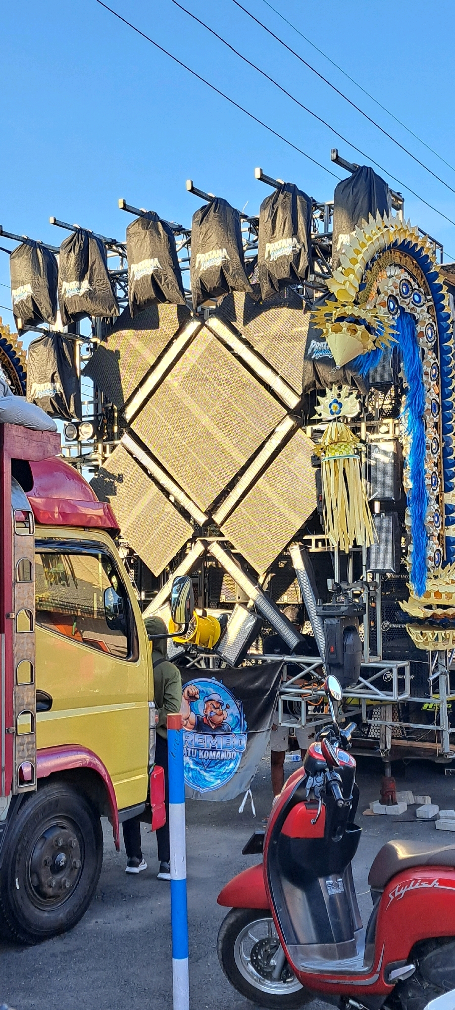 In Prigen, Indonesia, a yellow truck with a red canopy is parked next to a large, elaborate sound and lighting system. The sound system features a large, diamond-shaped LED screen and is adorned with a blue and gold dragon-like decoration on the right side. Several black fabric covers, labeled "PRATAMA Lighting," are draped over parts of the sound system. A banner featuring a cartoon character, possibly Popeye, with the text "REMBO ATU KOMANDO" is displayed on the front of the sound system structure. A person wearing a dark green hooded jacket stands near the truck, facing the sound system. In the foreground, a red scooter is parked. The scene is outdoors with a clear blue sky above, and power lines are visible stretching across the frame. The ground appears to be paved.