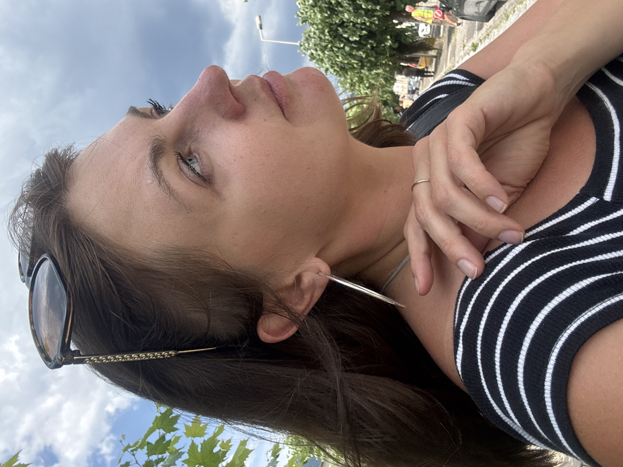 This media file features a close-up, low-angle shot of a woman's face and upper body, rotated 90 degrees counter-clockwise. She is looking upwards and to her left, with her eyes partially visible and her mouth slightly open. Her skin appears smooth with some freckles and a subtle sheen. A thin gold ring is visible on her right ring finger. She is wearing a black and white striped top, and a pair of sunglasses with a tortoiseshell pattern is resting on her hair, which is dark brown and falls around her shoulders. Long, thin earrings extend from her ears, one of which is clearly visible.
In the background, the scene is set outdoors. The sky is visible, with some white clouds against a blue backdrop. Green foliage, likely leaves from a tree or vine, is also present in the lower part of the frame. Further in the background, trees and what appears to be a street scene with people and vehicles can be seen, suggesting an urban or semi-urban environment. One person in the background is wearing a high-visibility yellow vest, possibly indicating an event or work crew. The lighting suggests it is daytime, likely late morning or afternoon, with bright, diffused light. The woman's expression is difficult to fully ascertain due to the angle, but it could be interpreted as contemplative or perhaps enjoying the moment.
No visible text is present in the media file. The overall impression is a candid, natural moment captured outdoors.