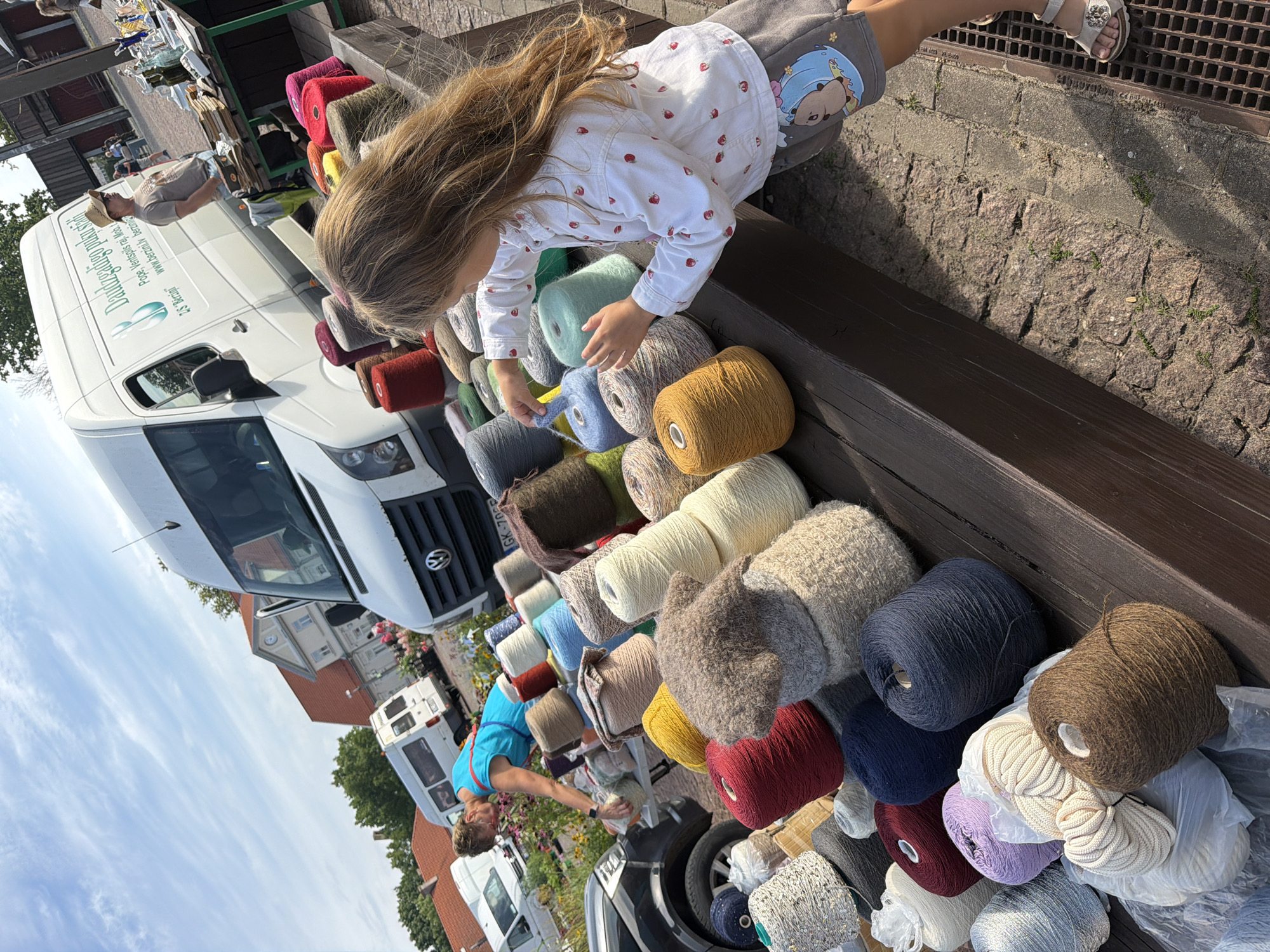 The image captures a vibrant outdoor market scene in Ventspils, Latvia, likely on a sunny day given the clear blue sky with scattered clouds. In the foreground, a young girl with long, blonde hair is intently examining spools of colorful yarn. She is wearing a white shirt adorned with small red strawberries and grey shorts with a cartoonish bear motif. Her hand reaches out, touching a light blue, fluffy spool of yarn, suggesting she is selecting or interacting with the materials.
Behind the girl, a wide array of yarn spools are displayed on a wooden table. These spools vary in size, texture, and color, ranging from earthy tones like brown, beige, and cream to bright reds, yellows, and blues, as well as muted greens and purples. Some spools are tightly wound, while others appear more loosely spun. In the background, a white van with the "Daudzra’dzigo pūkainu stāstu" logo and "DS 'Berzini'" text is parked. This van likely belongs to a vendor at the market. A woman in a bright blue top is visible near the van, appearing to be attending to the market stall or interacting with customers. Further in the background, more vans and market stalls are present, along with a few other individuals, indicating a bustling event. The environment suggests an open-air market with buildings and trees visible behind the stalls, typical of a town or city setting. The ground is paved with cobblestones.