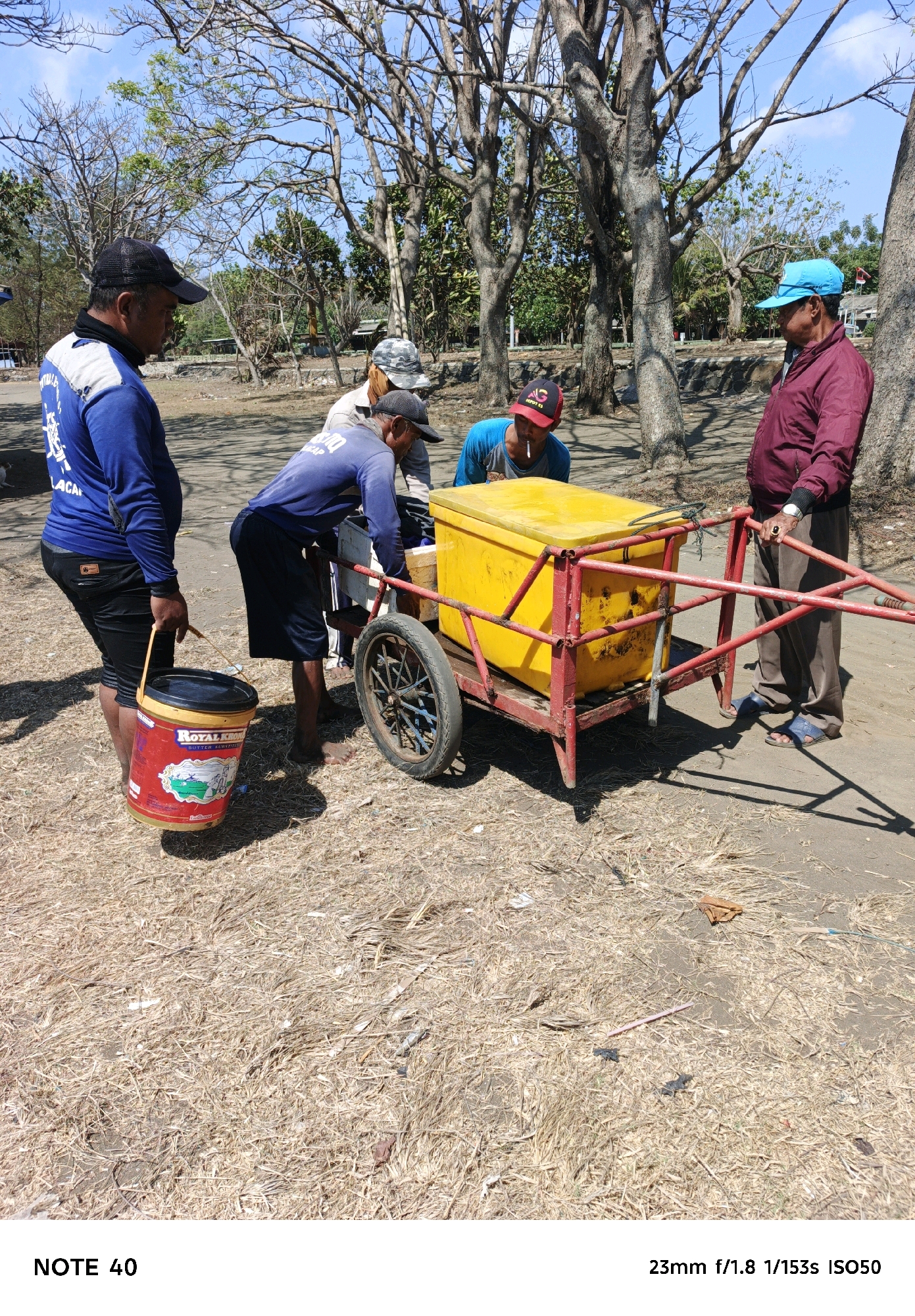 Four adult males are standing around a red and yellow cart loaded with a yellow cooler. The men are of Indonesian descent, and they are wearing casual clothing. One man on the left is wearing a blue long-sleeved shirt and black pants, and he is holding a red bucket. The second man from the left is wearing a blue long-sleeved shirt and black shorts, and he is smoking a cigarette. The third man from the left is wearing a light-colored shirt and camouflage hat. The fourth man from the left is wearing a maroon jacket and brown pants, and he is wearing a blue cap. The cart is on a dirt path surrounded by trees and dry grass. The scene appears to be outdoors.