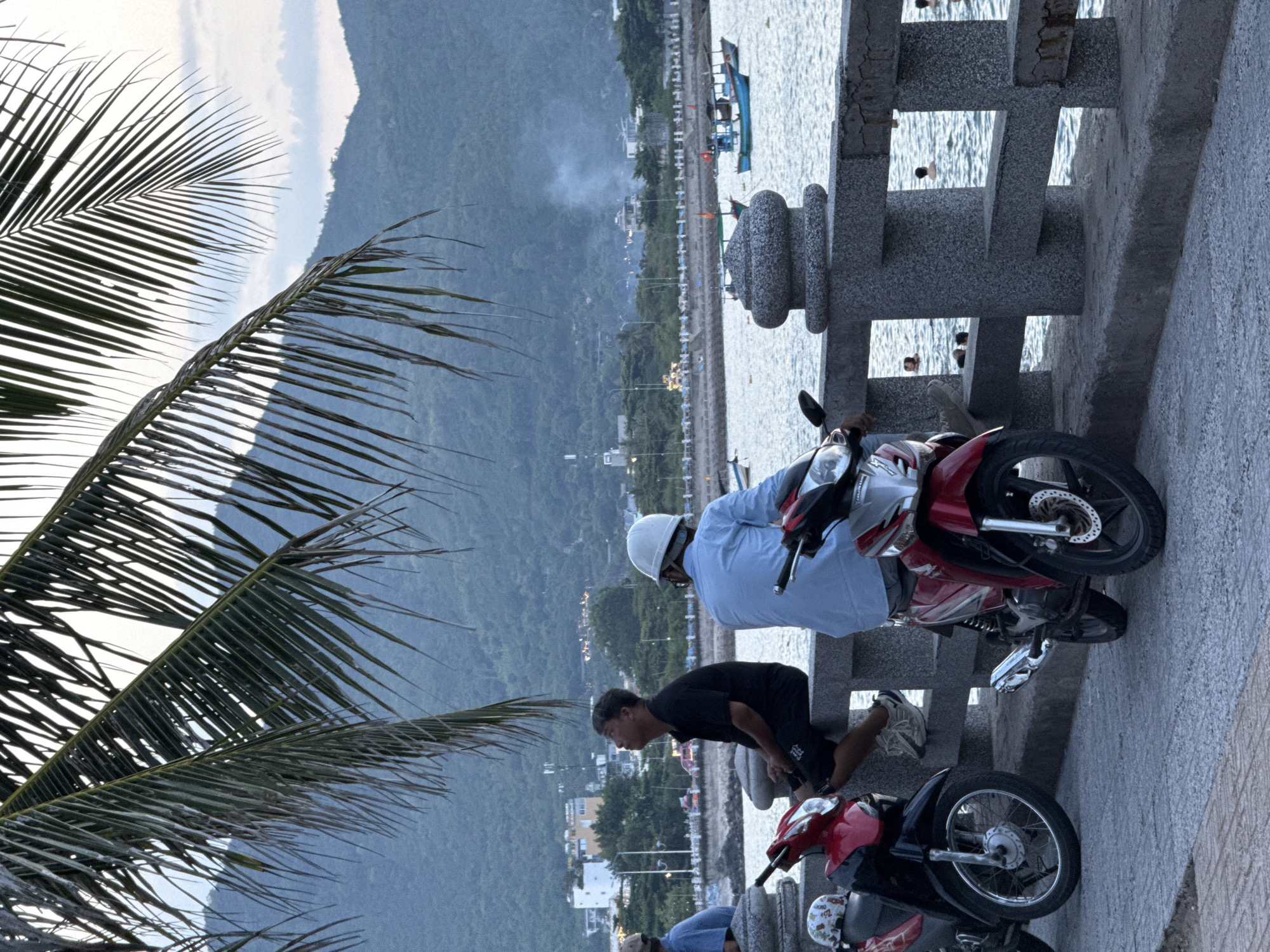 The image depicts a waterside promenade in Nha Trang, Vietnam, likely in the vicinity of Tran Phu Beach, given the presence of a scenic walkway, a body of water, and distant hills characteristic of the area. The time appears to be late afternoon or early evening, with soft, diffused light suggesting twilight.
In the foreground, two motorcycles are parked on a paved walkway. The one on the left is primarily red and black, while the one on the right is red and silver. To the left of these motorcycles, a person in a blue shirt is partially visible, seated. Adjacent to them, a man in a black shirt and dark shorts is seated on a stone wall overlooking the water. He is looking upwards. To the right of the seated man, another person, wearing a light blue shirt and a white hard hat, is seated on the same stone wall, facing away from the camera.
The stone wall forms a barrier between the walkway and the sea. The sea itself is calm, with several small boats visible in the distance, including a larger blue and white boat. In the background, a long, developed coastline is visible, with buildings and trees lining the shore. Beyond the coastline, lush green hills rise, their slopes covered in dense vegetation. The sky above is overcast with some lighter patches, indicating the transition to dusk. Several people are visible in the water, enjoying a swim or wading. Palm fronds from a nearby tree hang into the upper portion of the frame, framing the scene.
