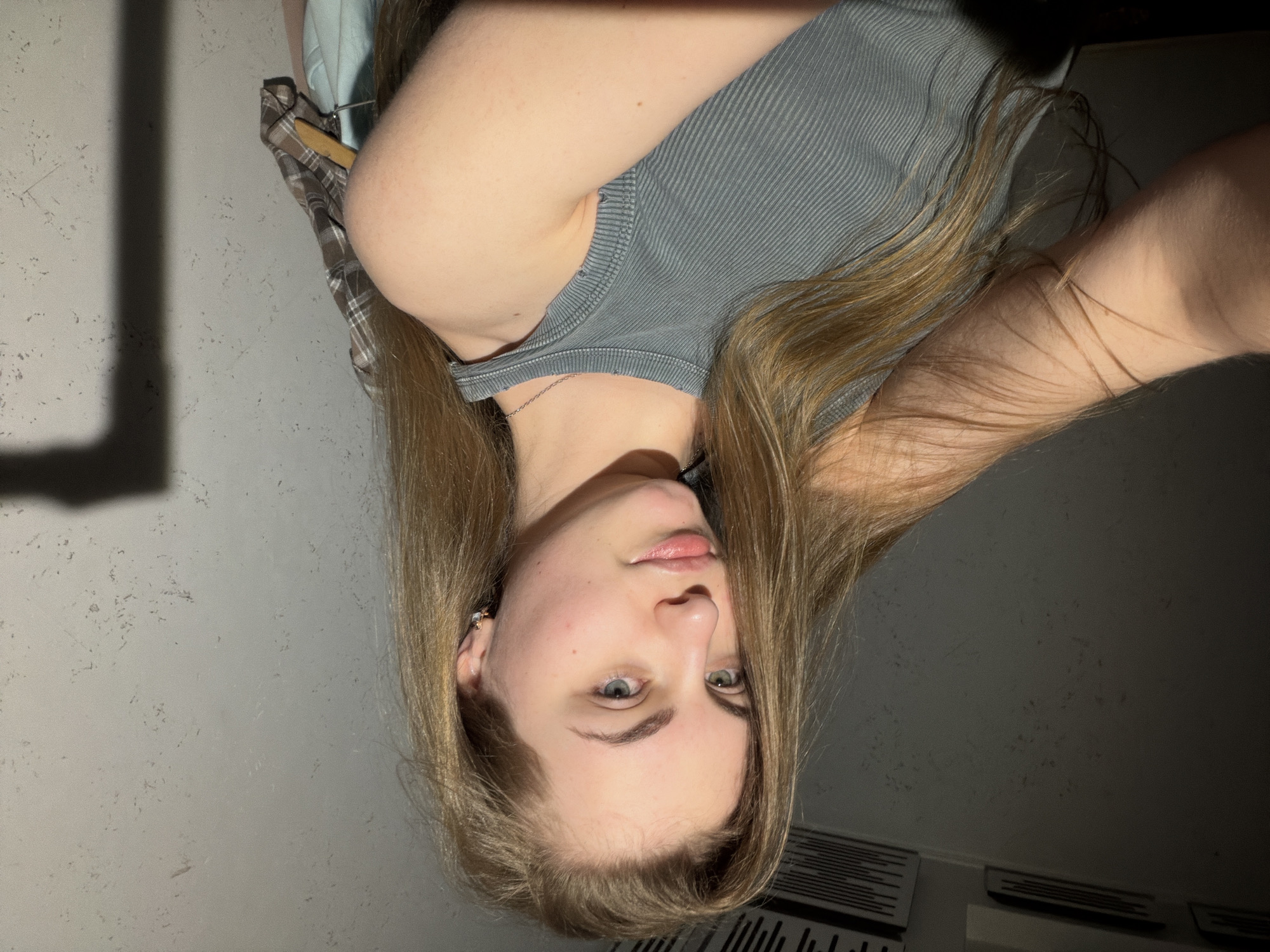 The image shows a young woman with blonde hair and light-colored eyes, looking towards the camera. She is wearing a gray ribbed tank top and a silver necklace. Her hair is long and styled down, framing her face. The background appears to be an indoor setting with a textured gray wall. To the left, there's a hint of plaid fabric and a wooden hanger, suggesting clothing might be nearby. The lighting is dim, casting shadows on the wall and creating a somber mood. The image appears to be a selfie taken in a private space, possibly a bedroom or dressing area. There is no visible text in the image, and the time of day and weather conditions are not discernible. The woman's expression is neutral, with her lips slightly parted.